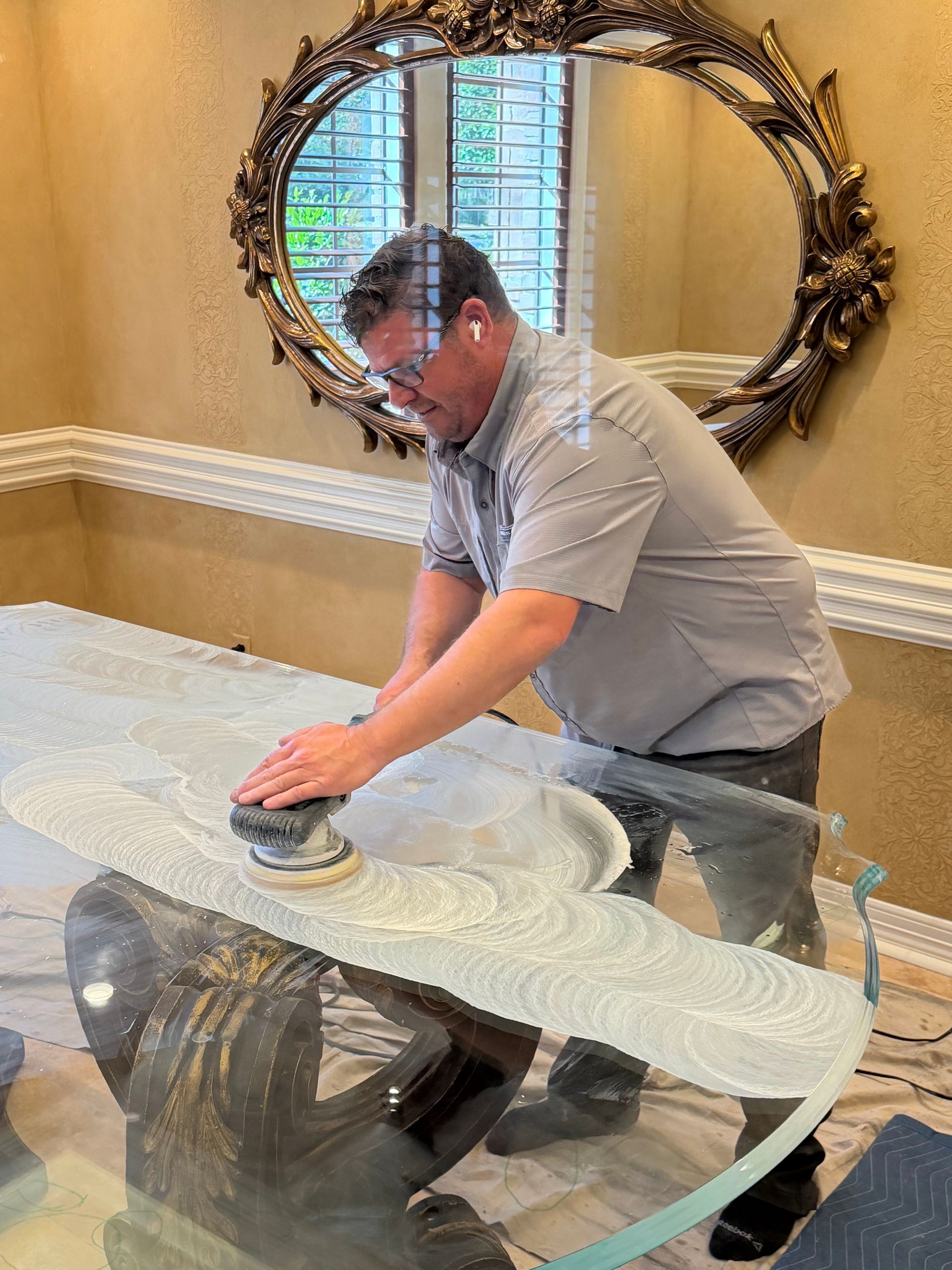 Man polishing a glass dining table with a cloth in a beige room with a decorative wall mirror