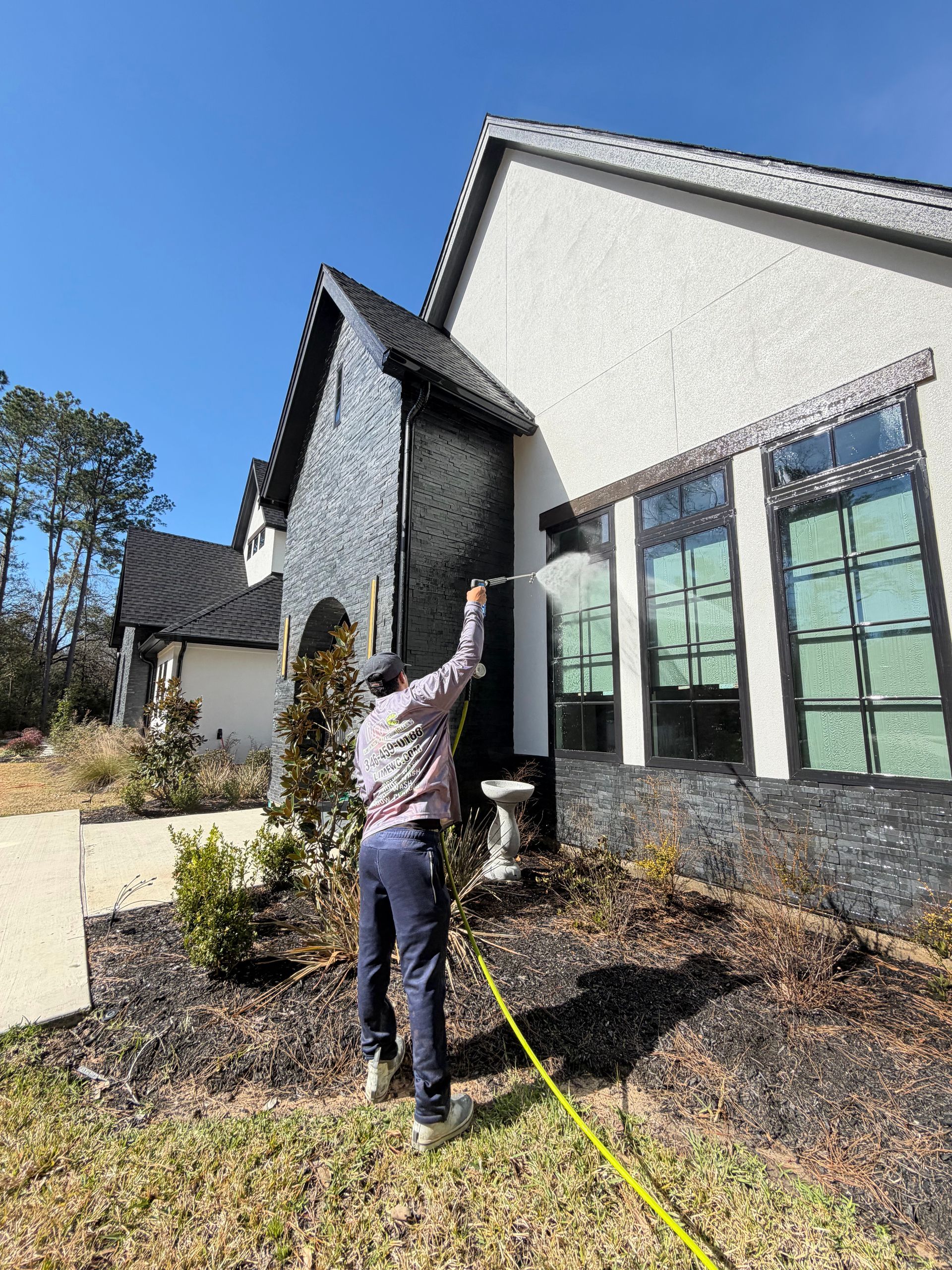 A person sprays water from a hose against the dark-tiled exterior wall of a modern home with white stucco siding.