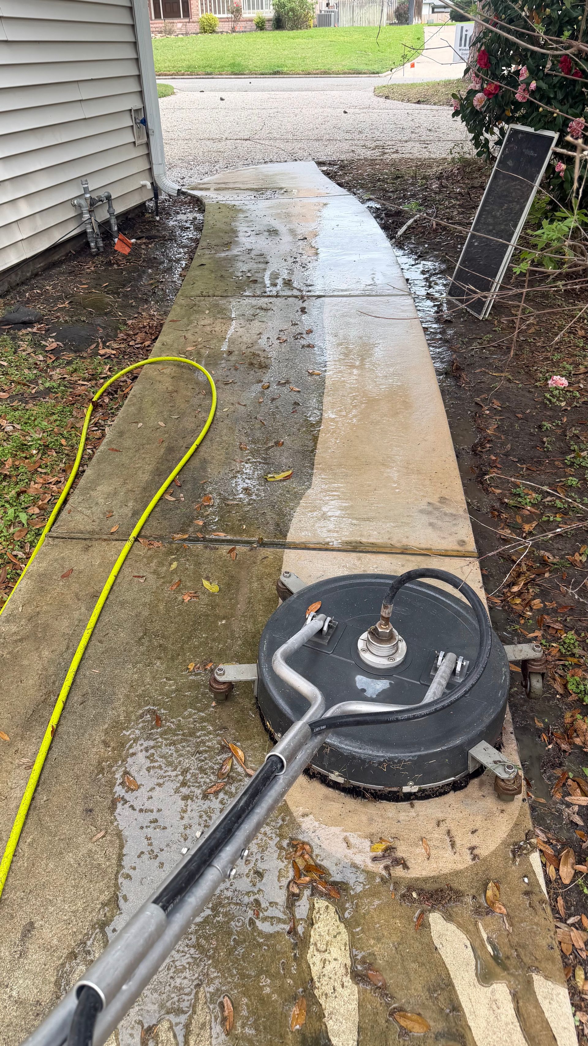 Driveway sidewalk being pressure washed, with a gray cleaner and a yellow hose along the house.