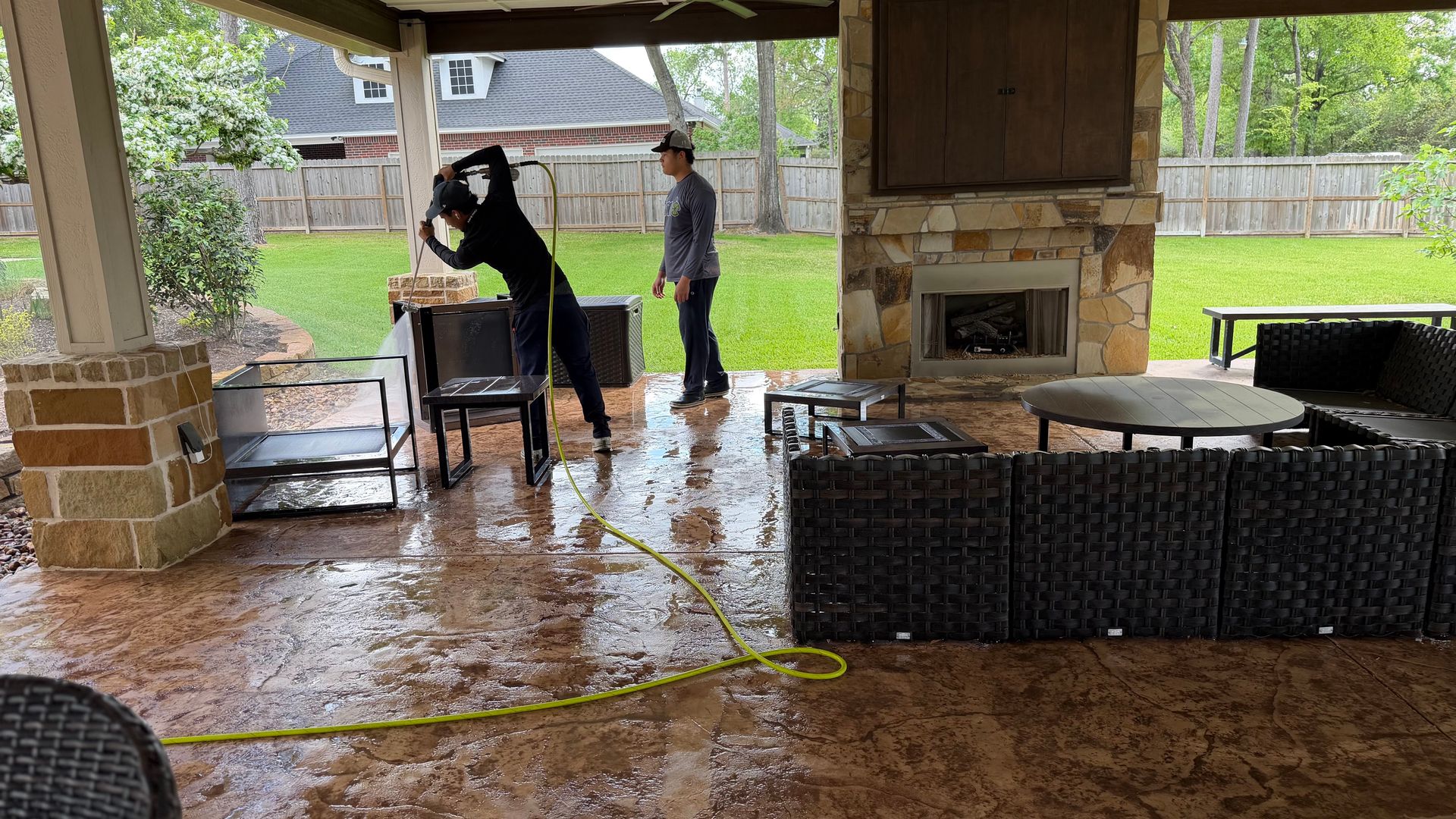 Covered patio with two people near a table, wet floor, and a grill in the background.