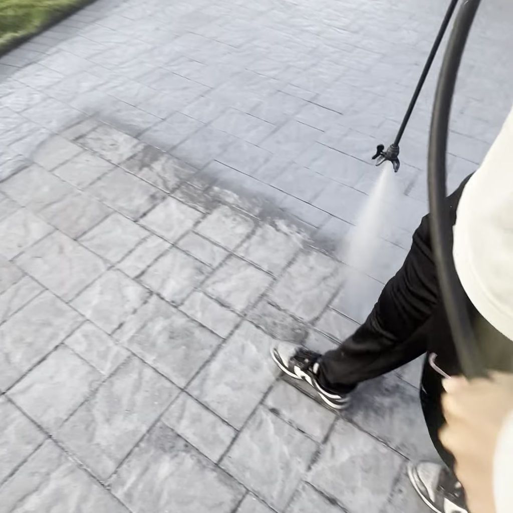 A person pressure washing a grey stone patio, cleaning a dark, dirty strip from the surface.