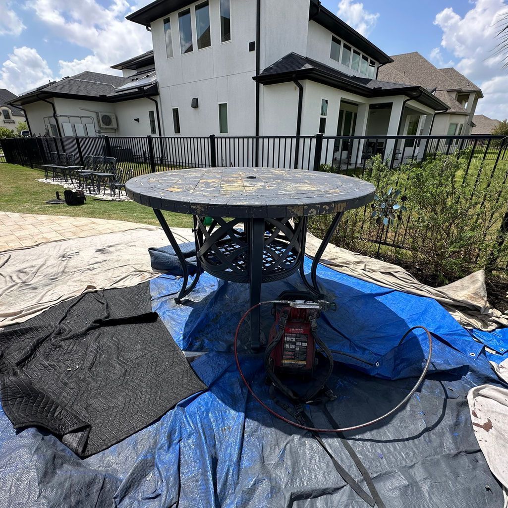 A black outdoor patio table sits on a blue tarp in a backyard, with a paint sprayer underneath.