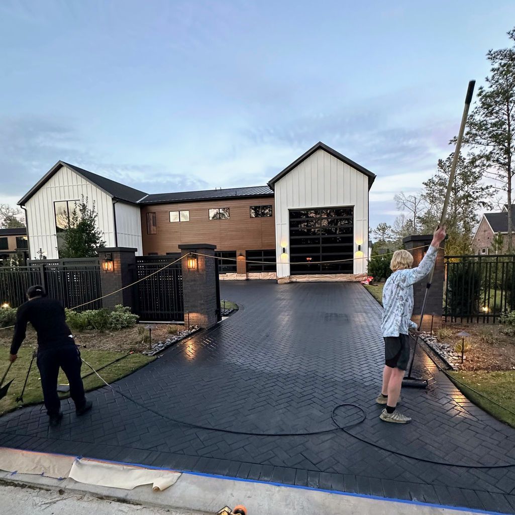 Two people use poles and hoses to apply a dark, textured coating to a residential driveway in front of a modern house.