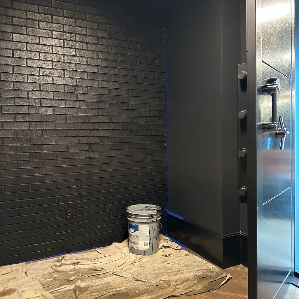 A black brick wall next to a dark painted pillar and a metal door, with a paint bucket on a drop cloth.