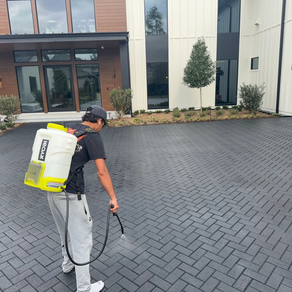 A worker uses a backpack sprayer to treat a dark, herringbone-patterned paver driveway in front of a modern home.