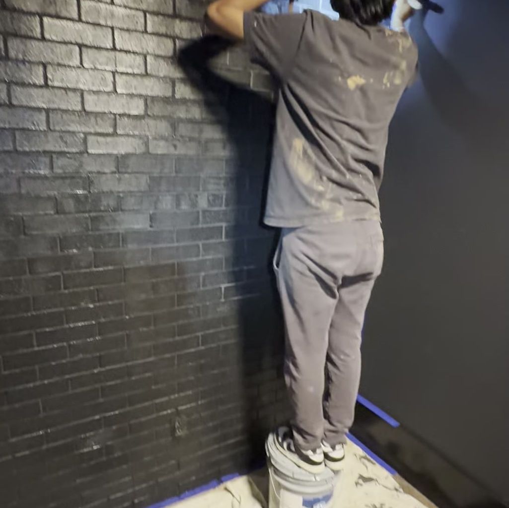 A person standing on a paint bucket, painting a dark brick wall with a roller.