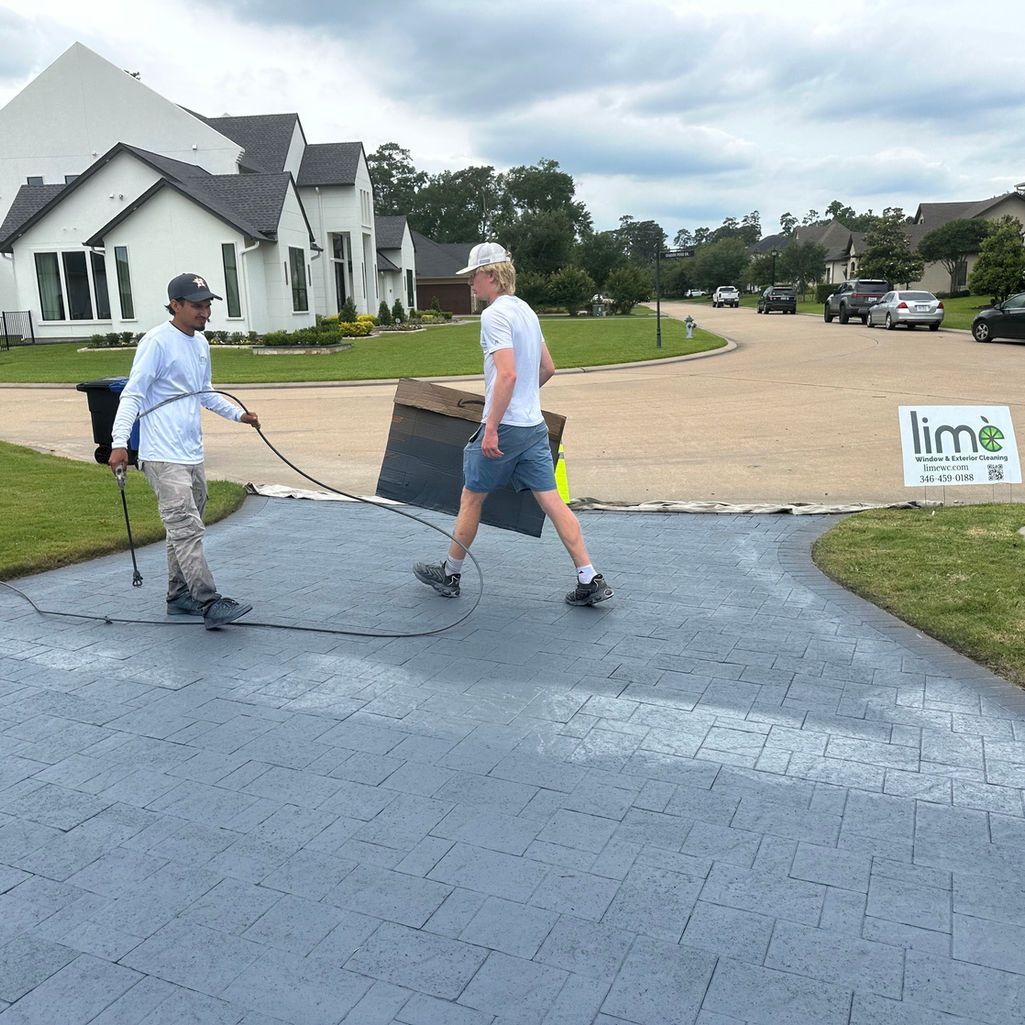 Two people work on a gray, patterned driveway in a residential neighborhood, one holding a tool and one carrying a mat.