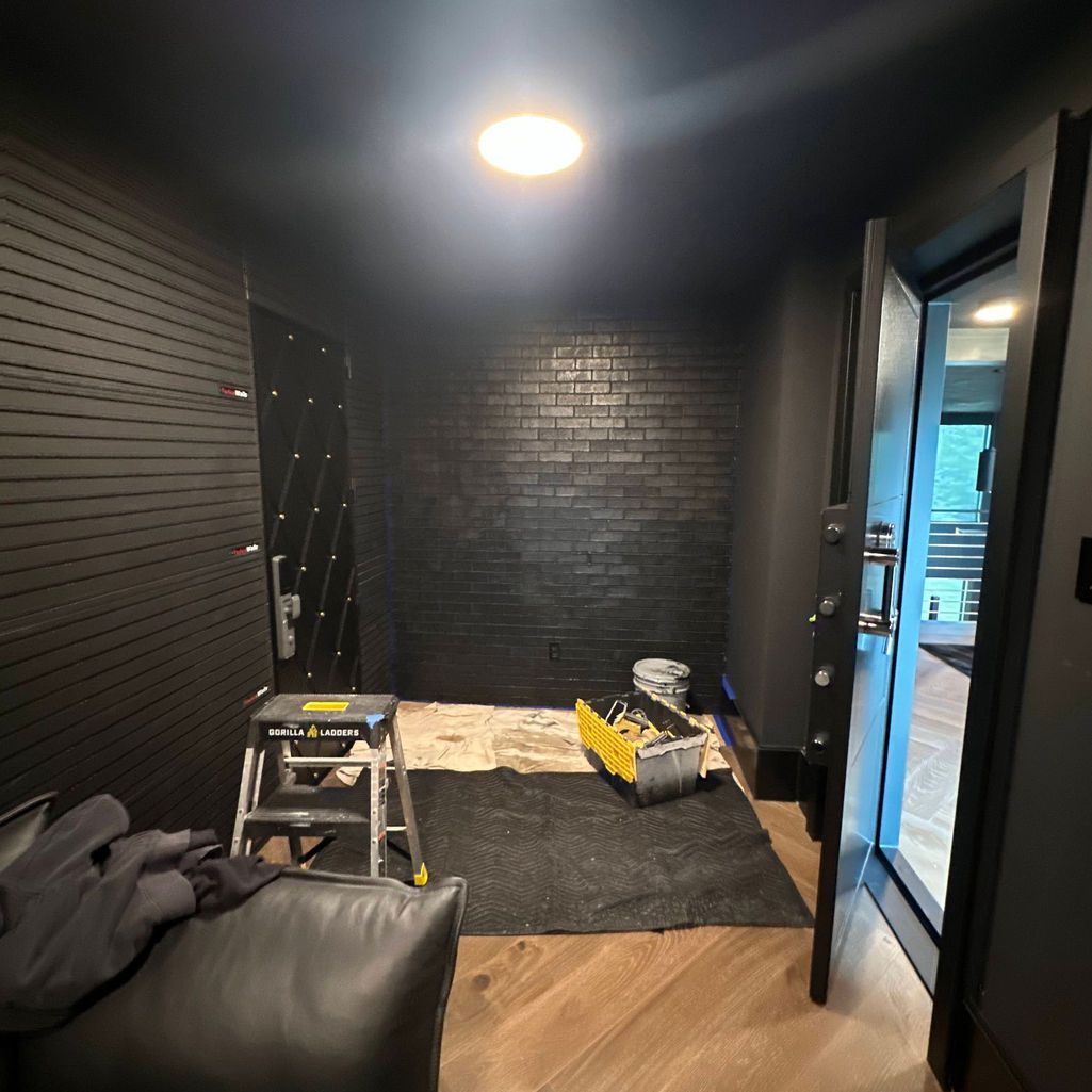 A renovation project in a small room with black walls, a brick accent wall, hardwood floors, a stepladder, and a bin.