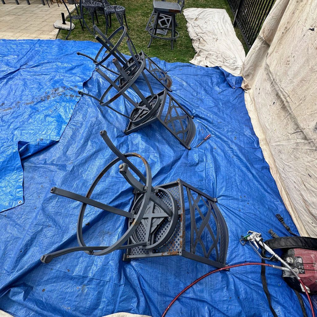 Two dark-colored metal chairs sit upside down on a blue tarp outdoors, presumably being prepped for painting.