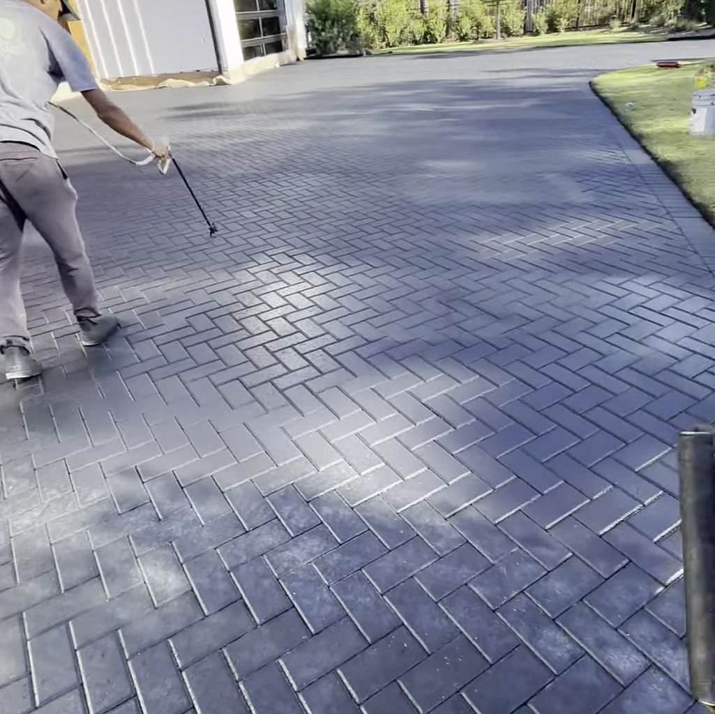 A person sprays a sealant onto a grey, herringbone-patterned brick driveway.