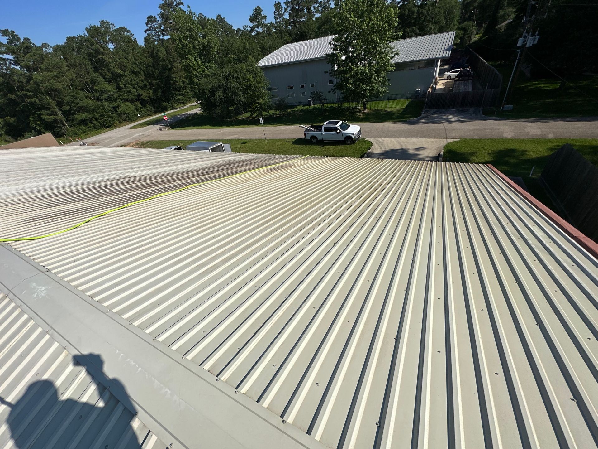 Ribbed metal roof with a shadowed person in the foreground and a white vehicle near a wooded house in back