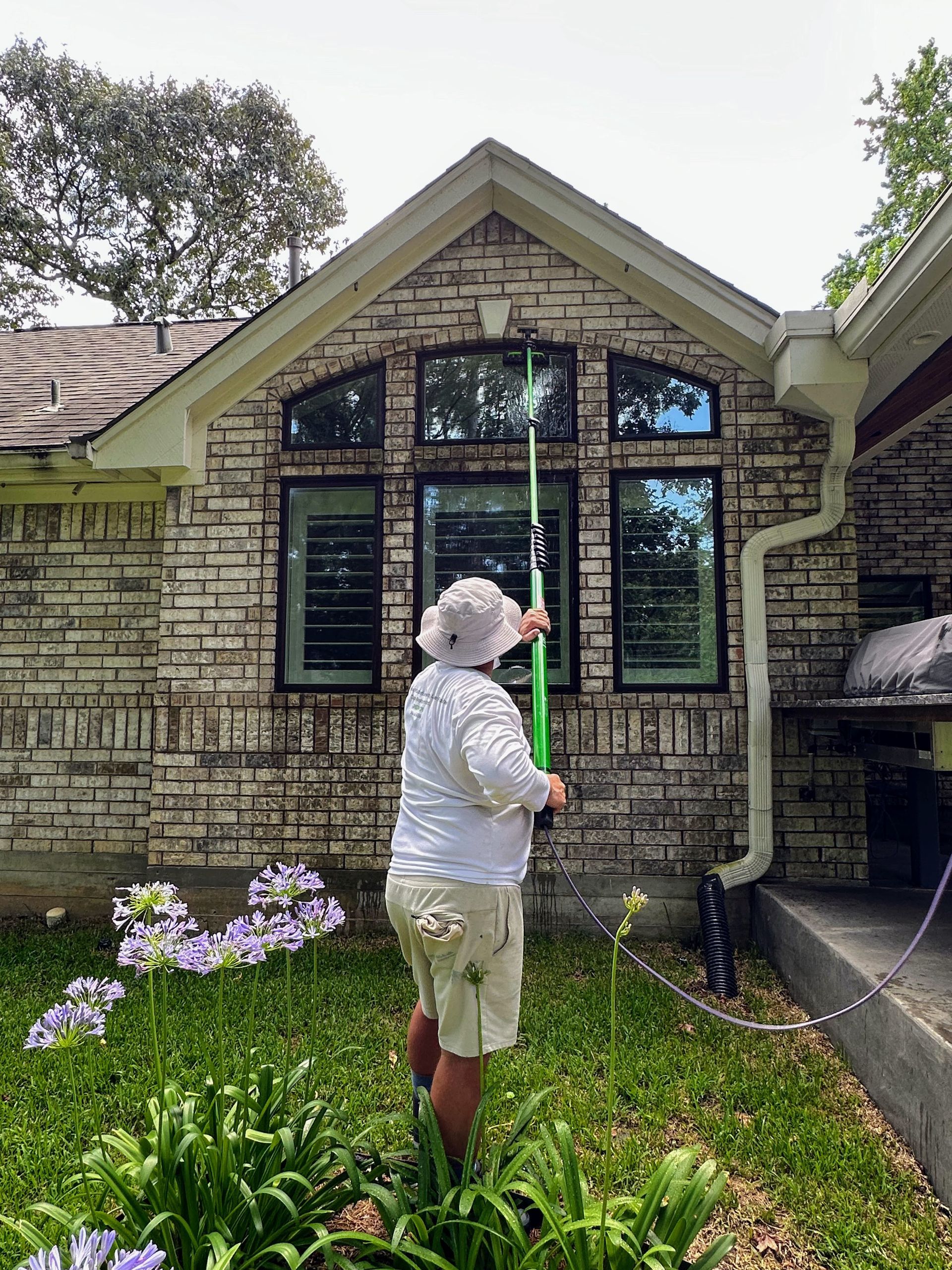 Person in a white hat inspecting a house window and trim in a flowered yard