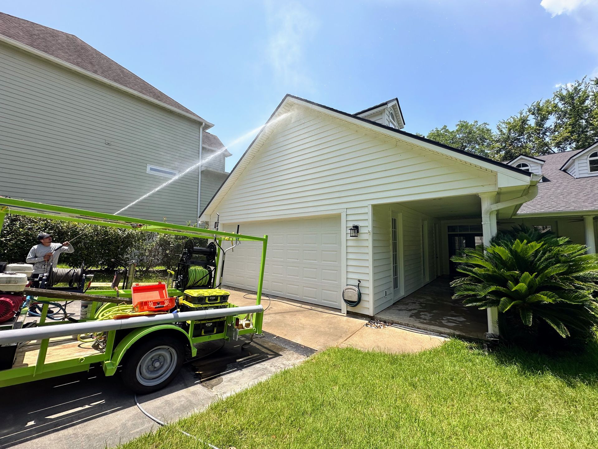 Worker pressure-washes a white house exterior beside a trailer and green lawn