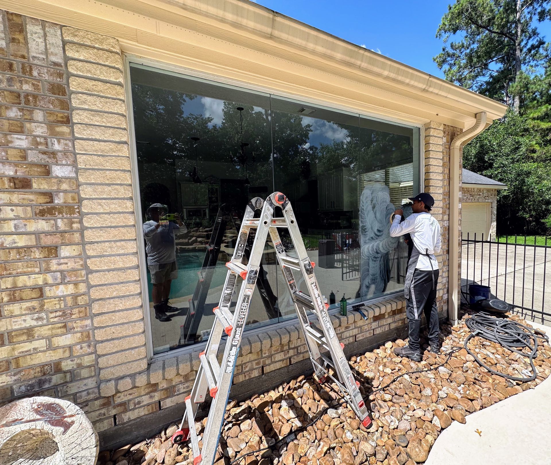 Workers installing a large window in a brick house exterior, with ladders and construction debris nearby