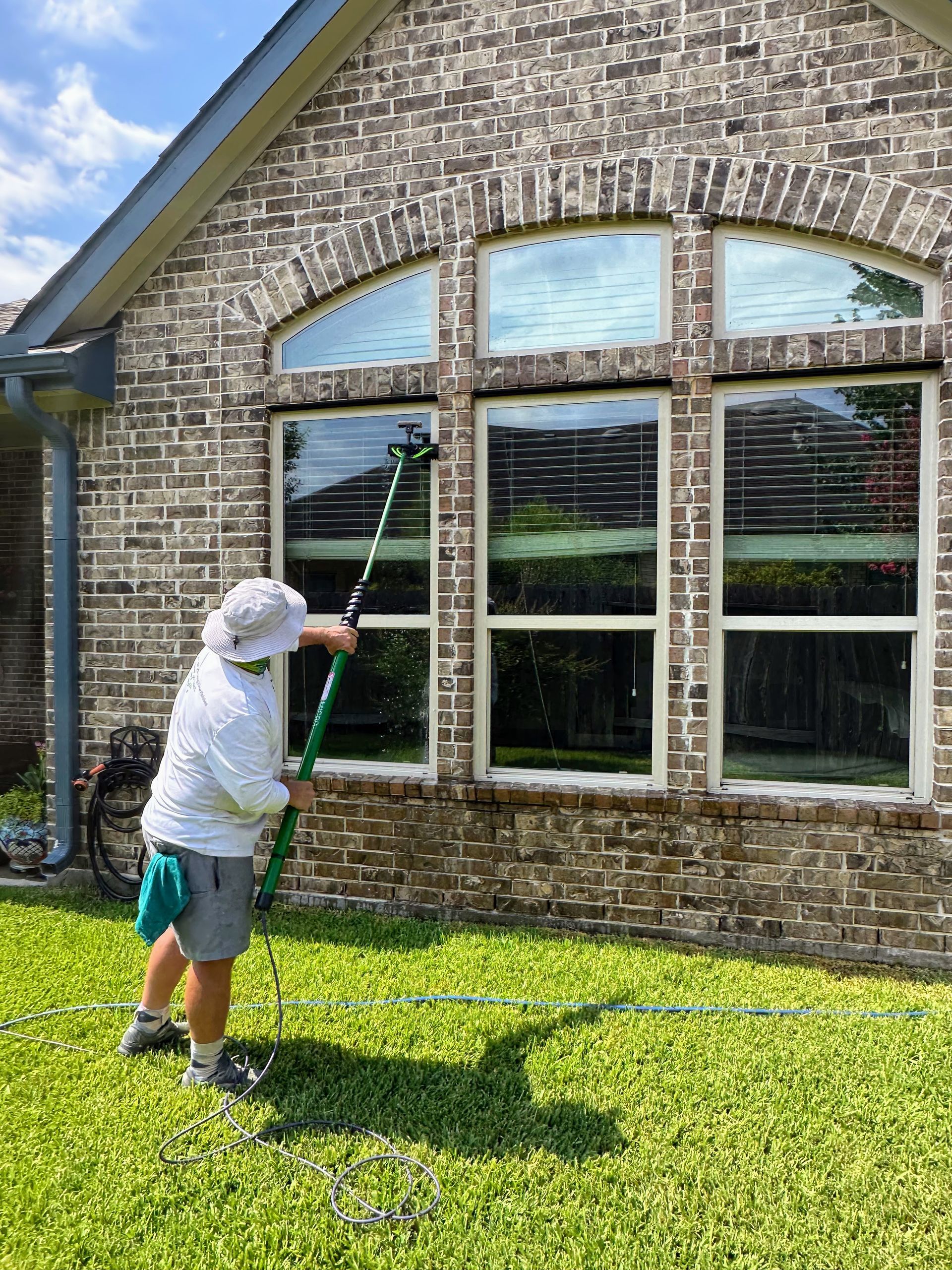 Child cleaning large brick house windows with a green squeegee on a sunny lawn