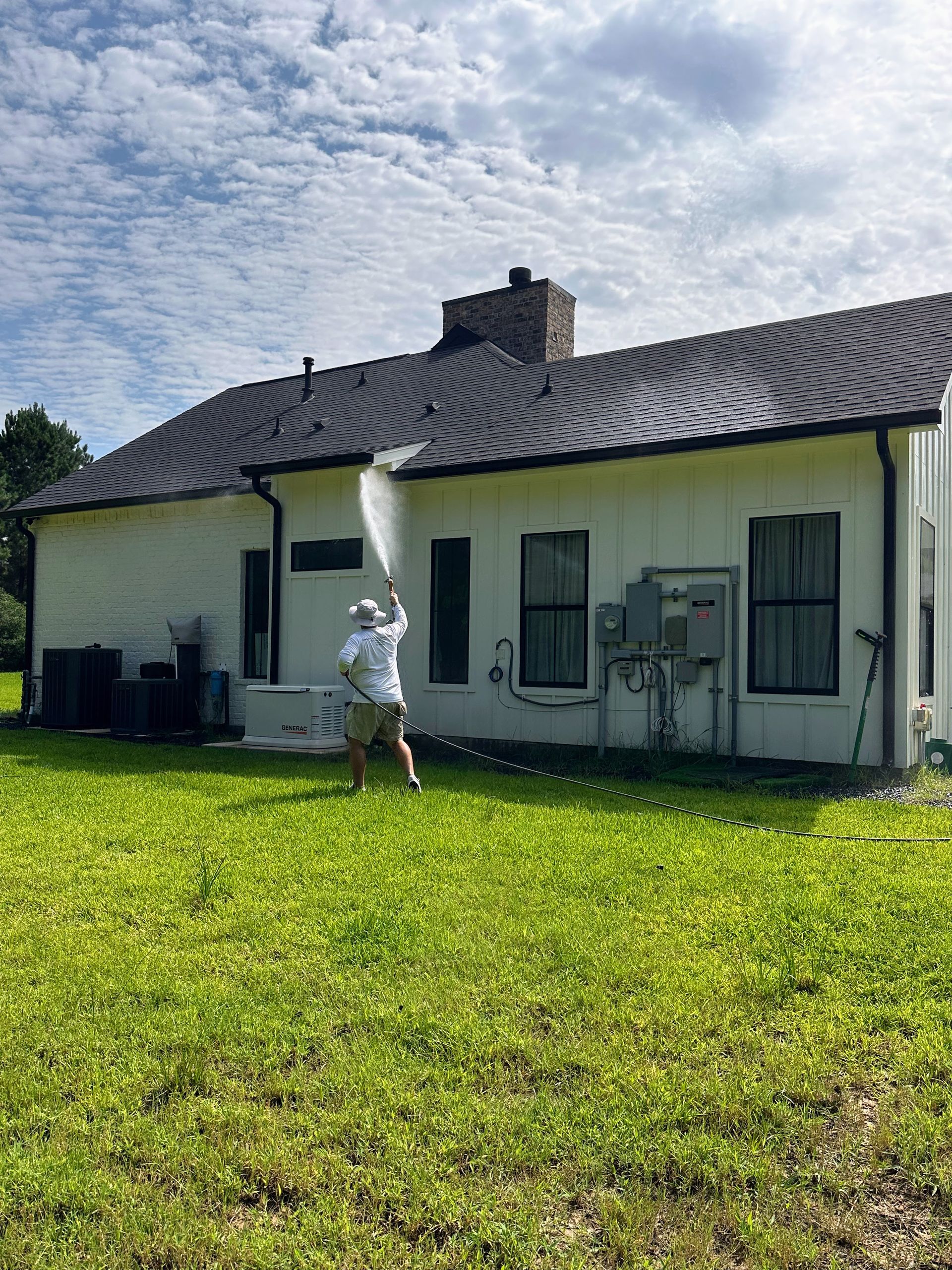 Person in white suit standing in a grassy yard beside a white house with a dark roof on a sunny day