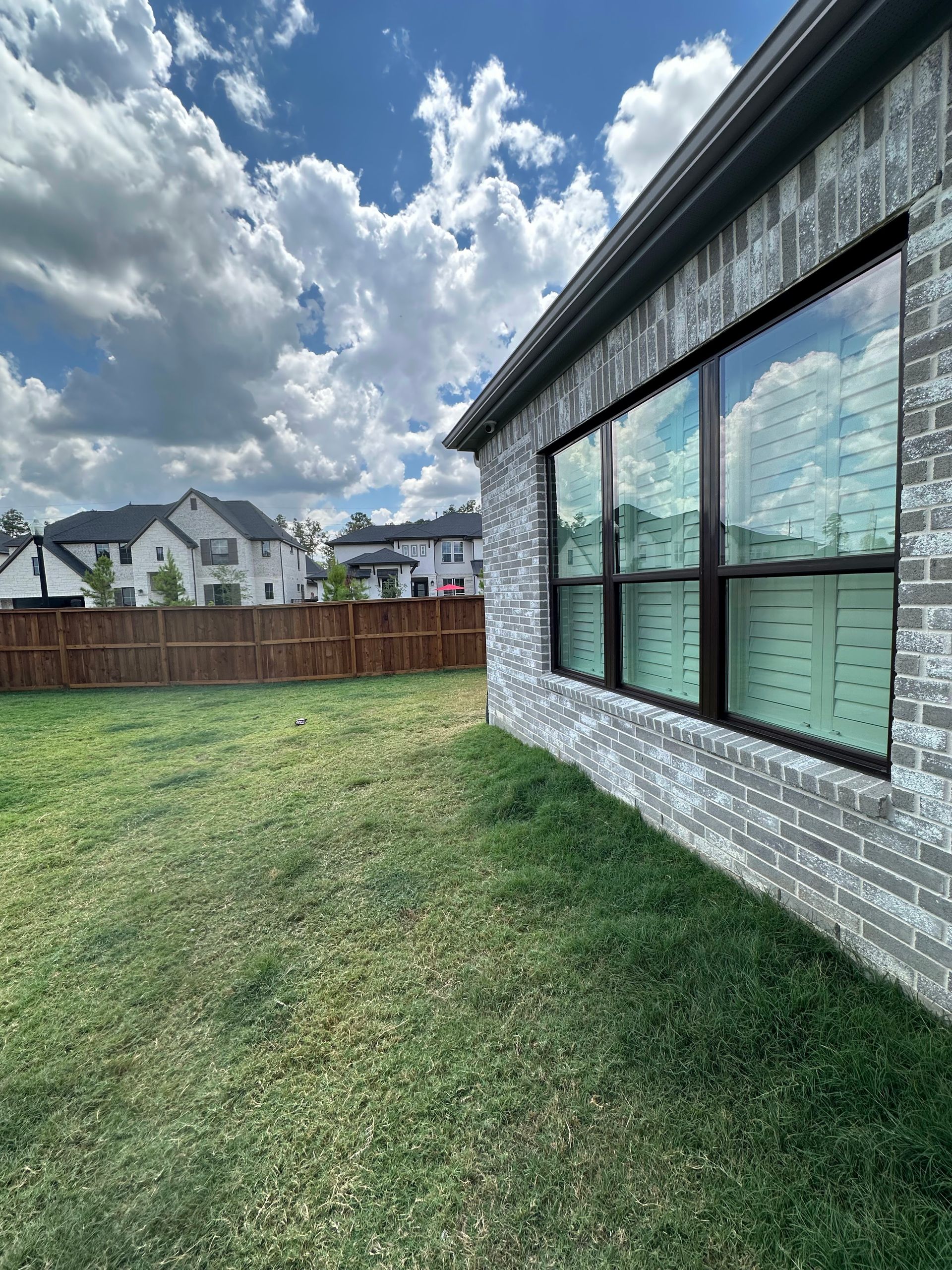 Backyard with green lawn, wooden fence, neighboring houses, and large windows on a brick home under a cloudy sky