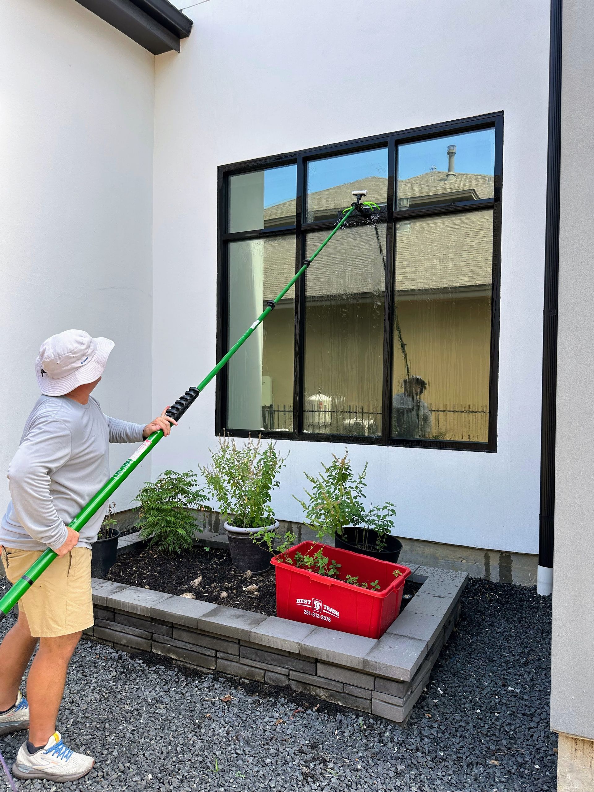 Person using a long green pole to clean a window beside a small garden and red crate.