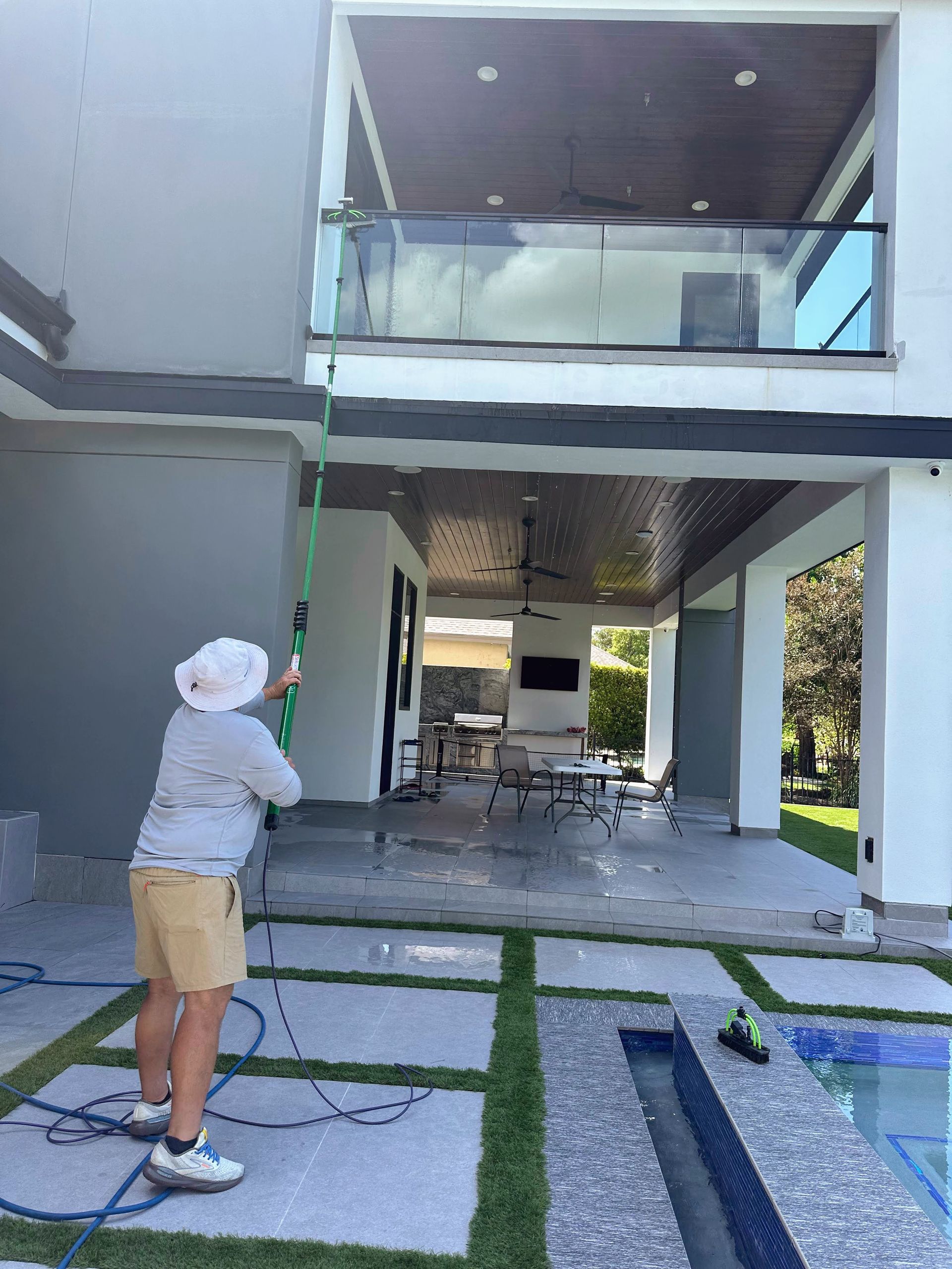 Person in a white hat standing in a modern courtyard, holding a hose and spraying water toward an open balcony.