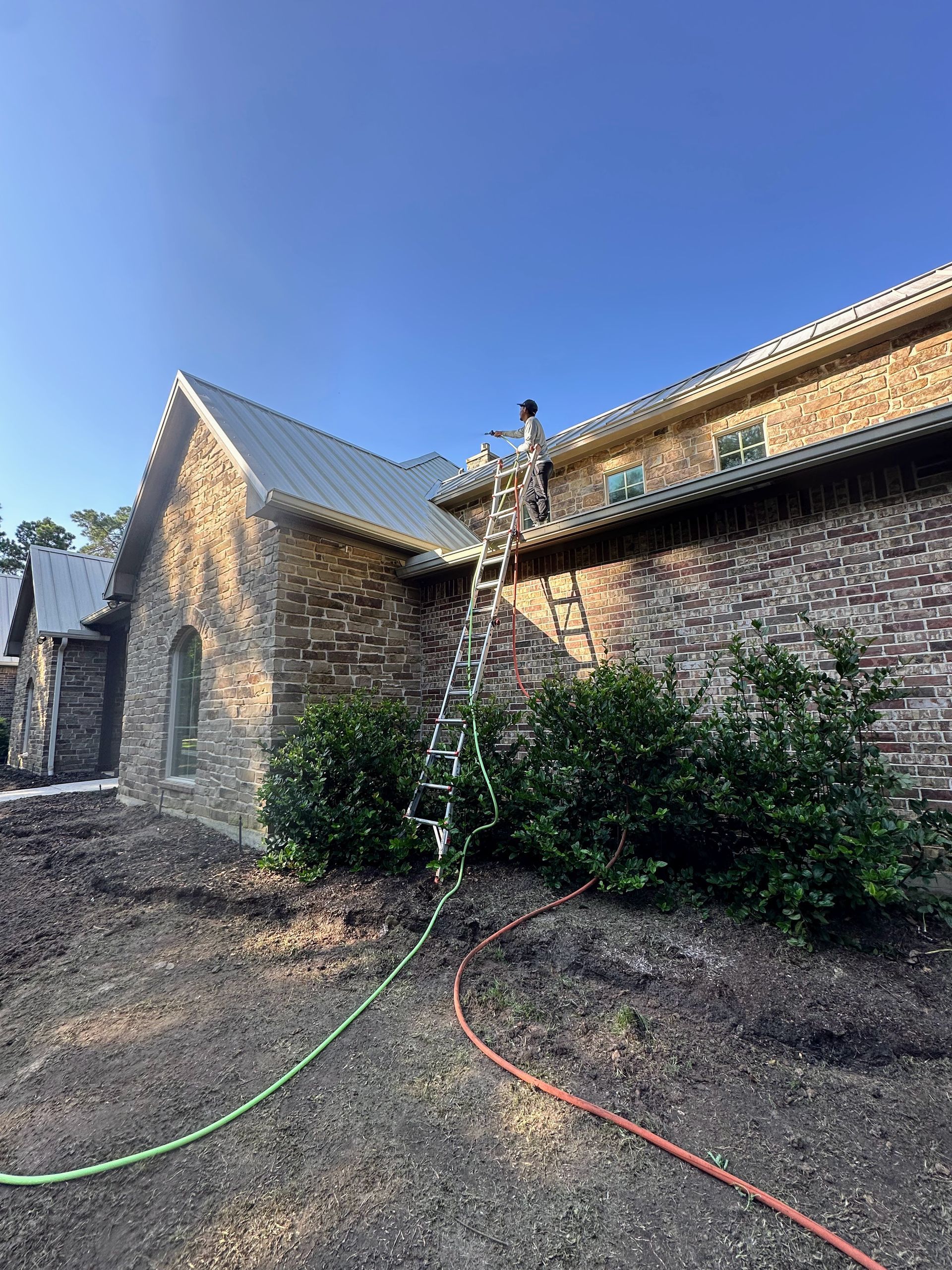 Worker on ladder cleaning brick house roof with hose in a yard under a blue sky