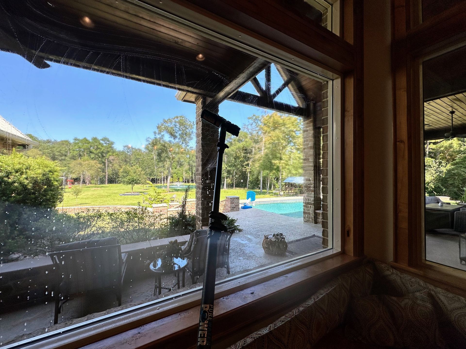 Backyard pool and patio viewed through a window, with trees and a sunny lawn beyond