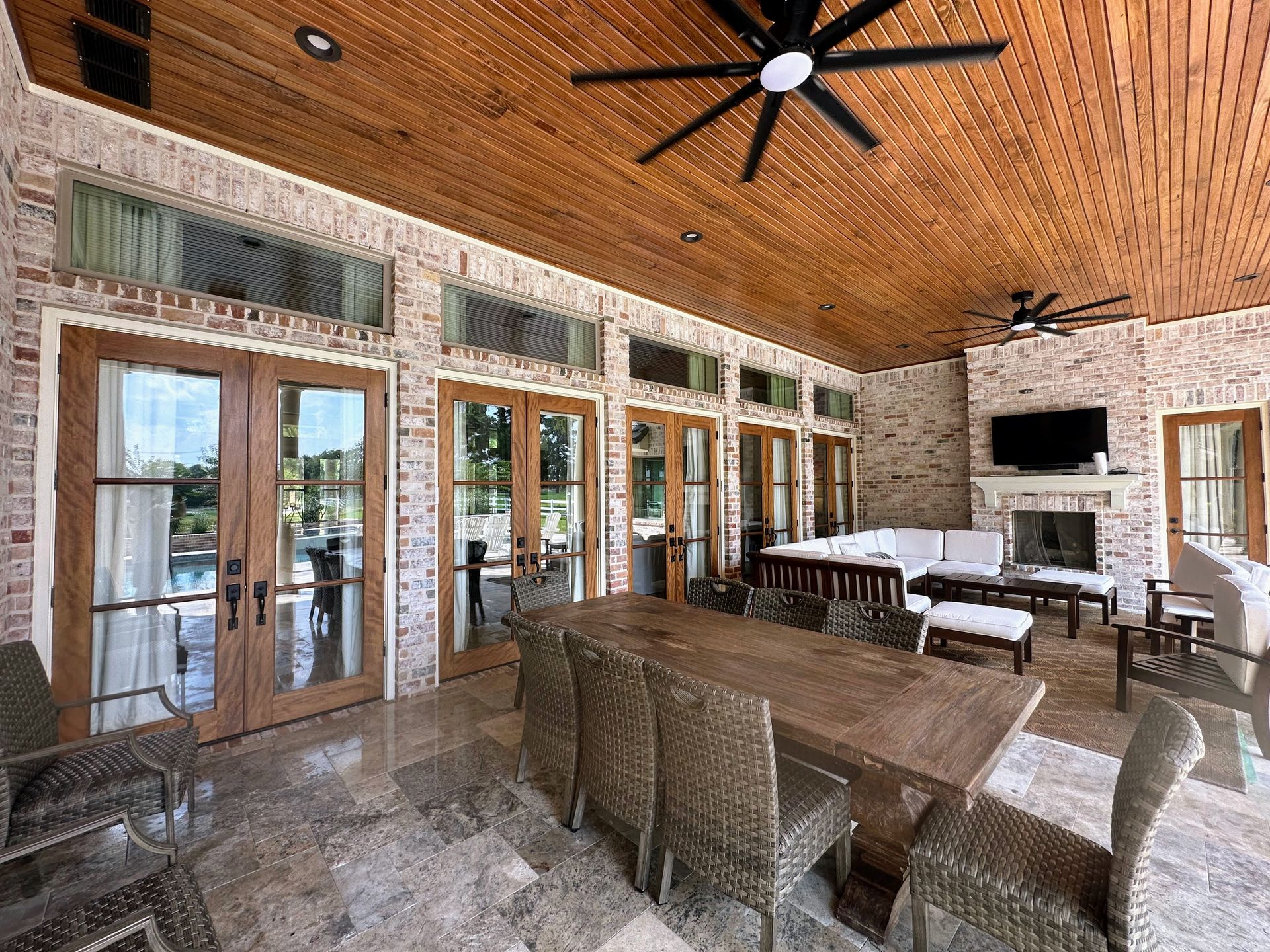 Covered patio with wicker seating, wood ceiling fans, glass doors, and a stone fireplace by the water