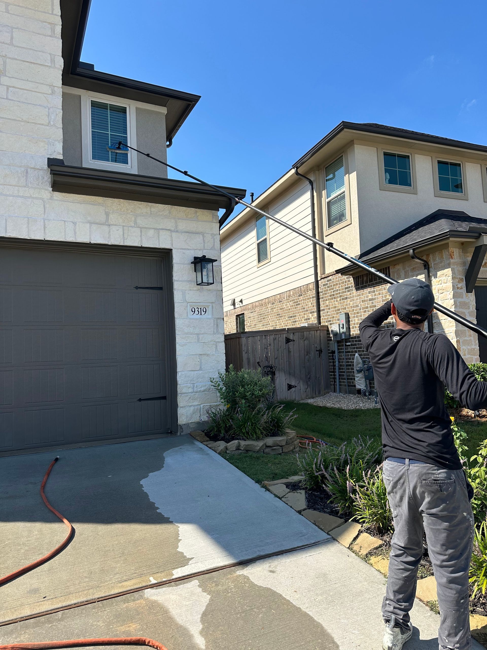 Person standing in a driveway beside a garage, aiming a hose toward a house exterior.