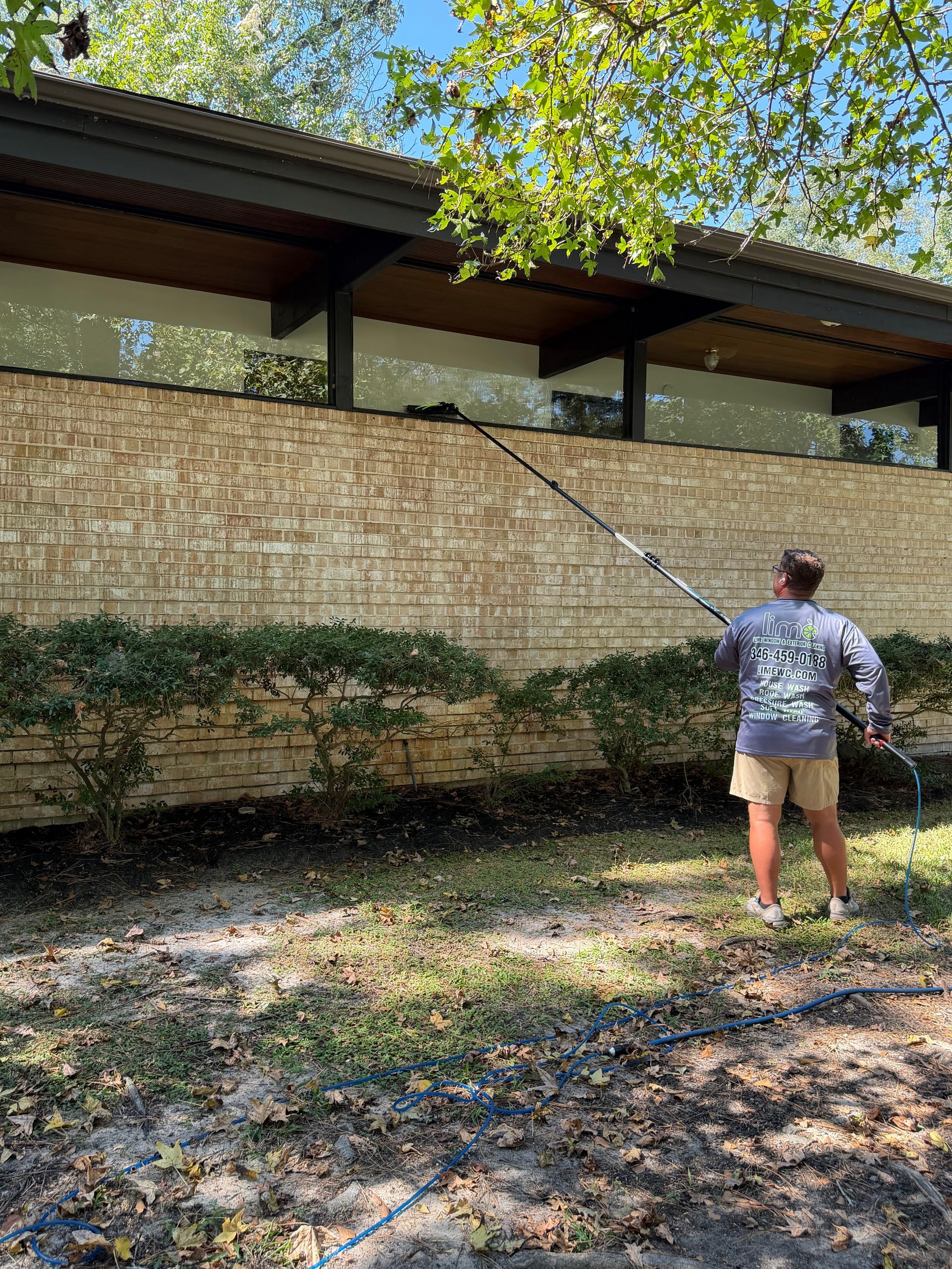 Person pressure-washing a brick wall beside a house under trees.