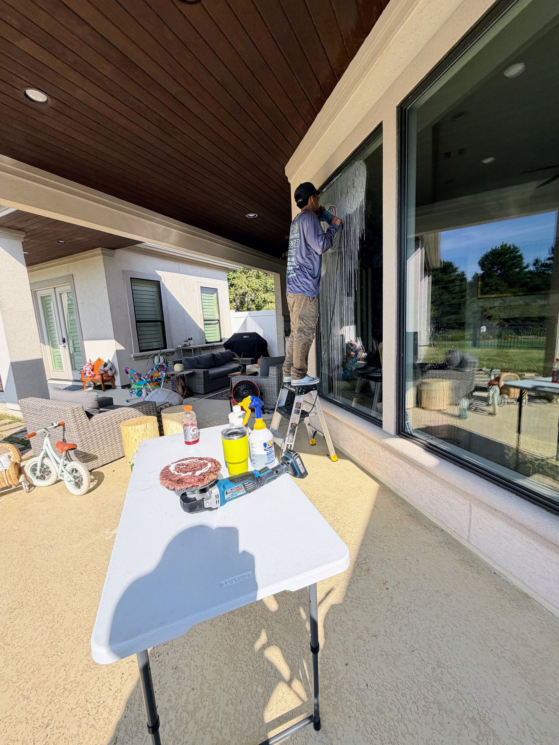 Person cleaning a large window on a covered patio beside a white table and scattered cleaning supplies
