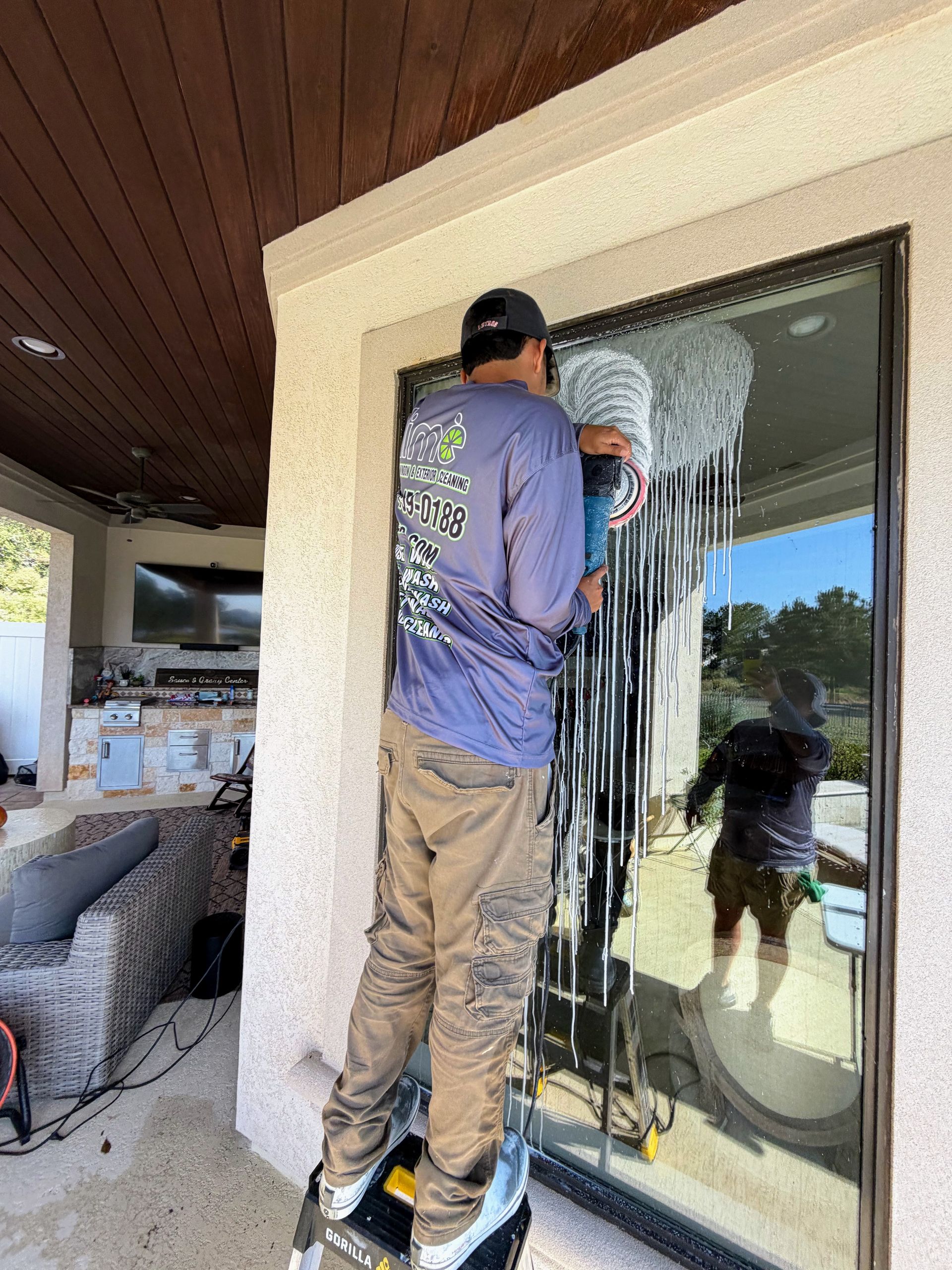 A person standing on a ladder cleans a large exterior glass door on a patio, with a reflection of the area visible.