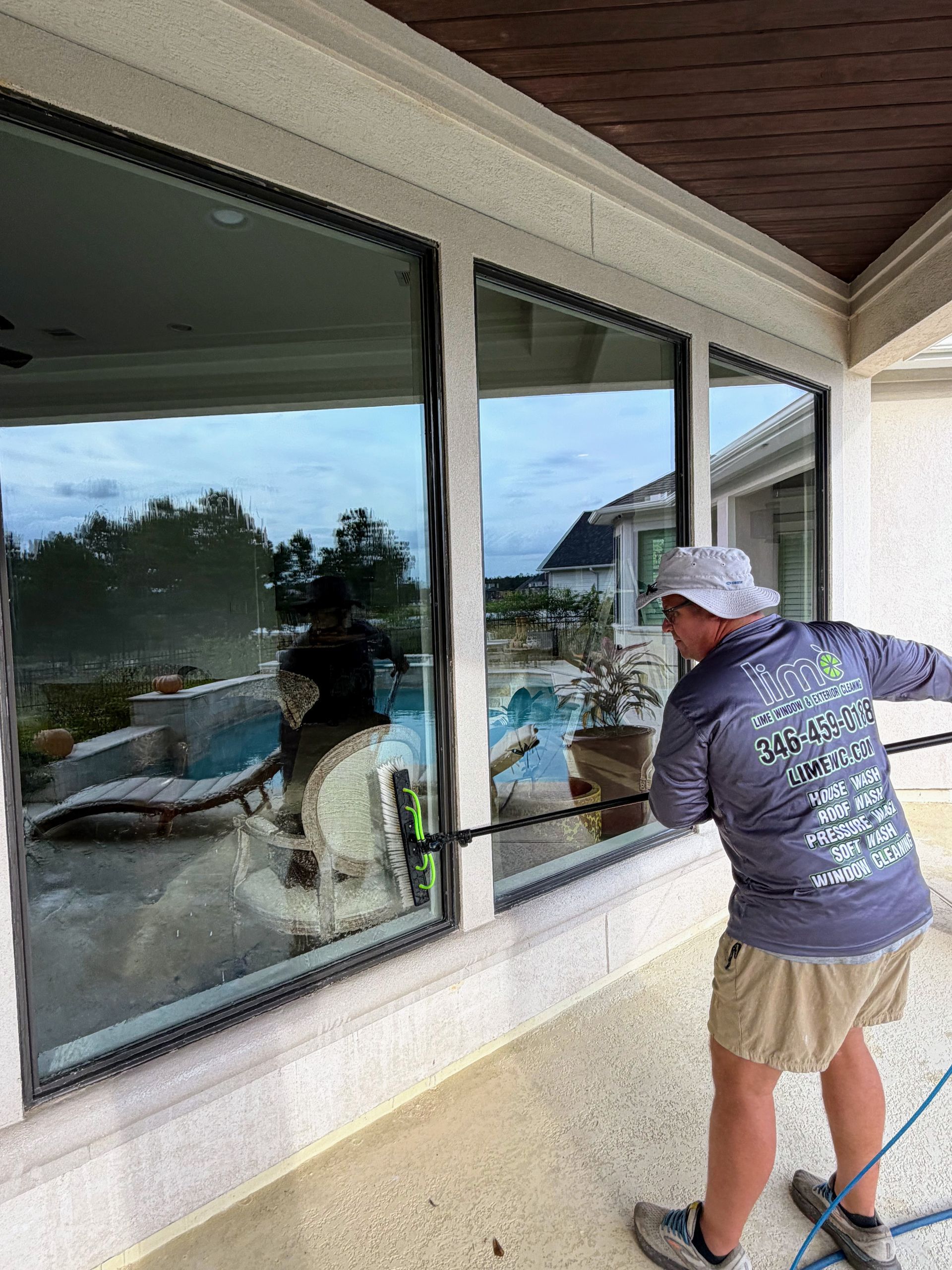 Person washing exterior windows of a house on a patio, with a pool reflected in the glass.