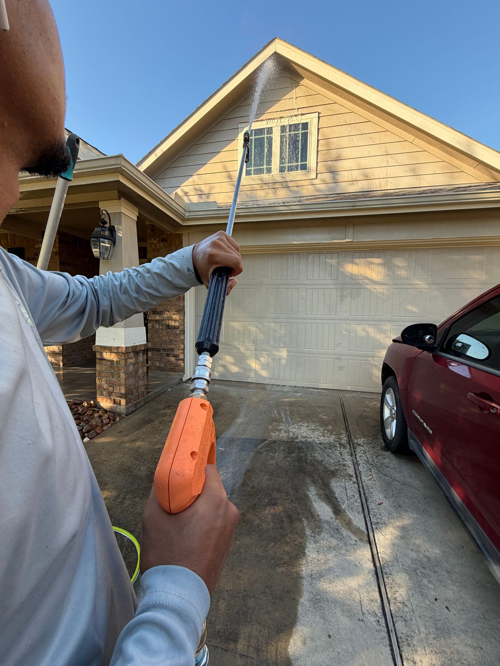 Person using a power washer on a driveway beside a red car and garage in a suburban driveway