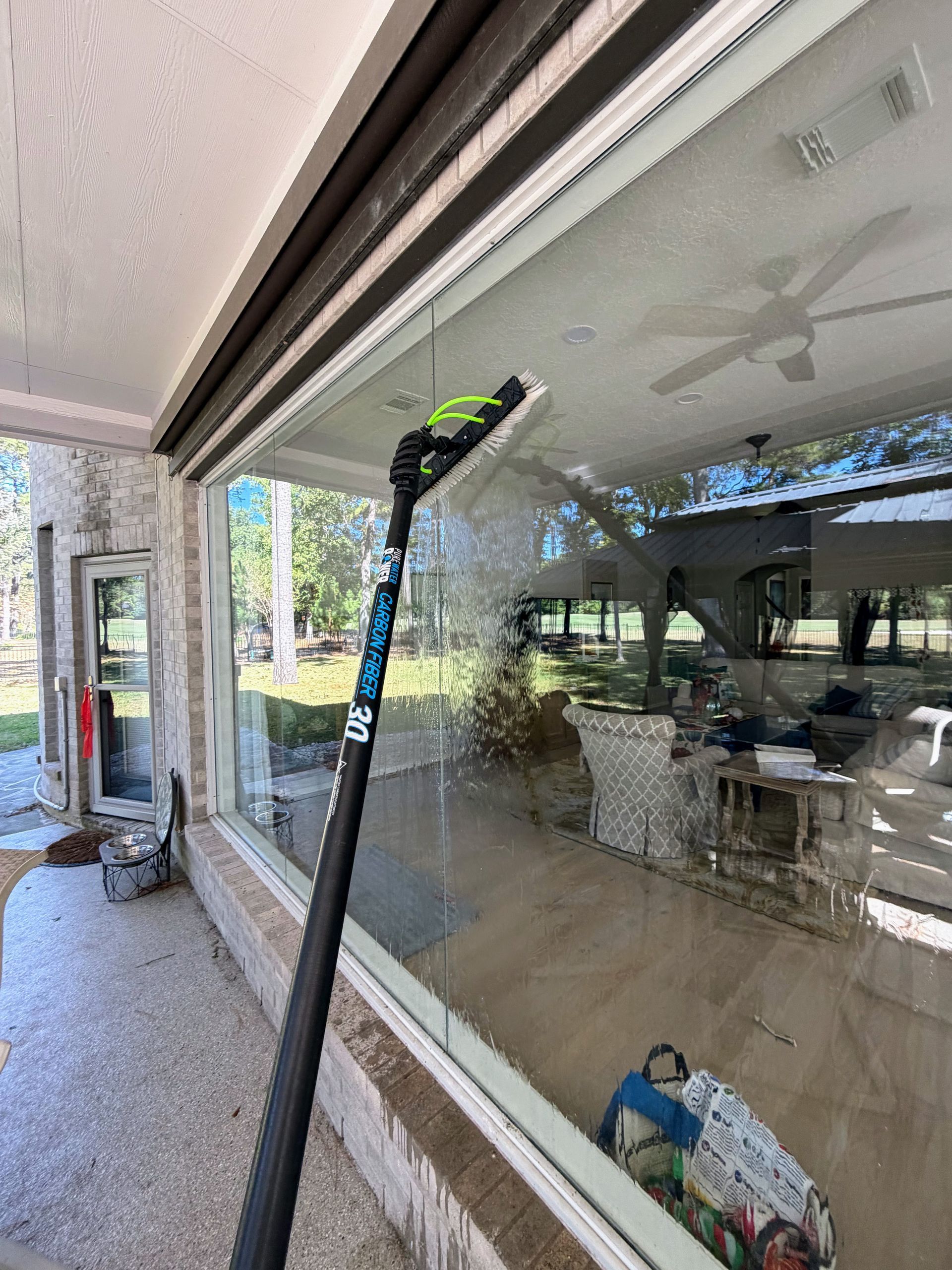 Long pole cleaning a glass patio enclosure with outdoor seating reflected in the windows.