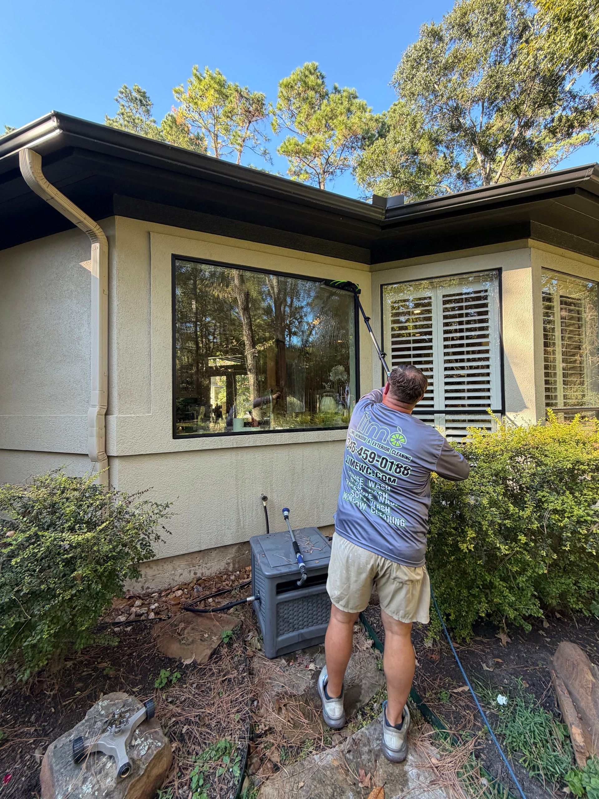 Person cleaning an exterior window on a beige house with bushes and trees nearby