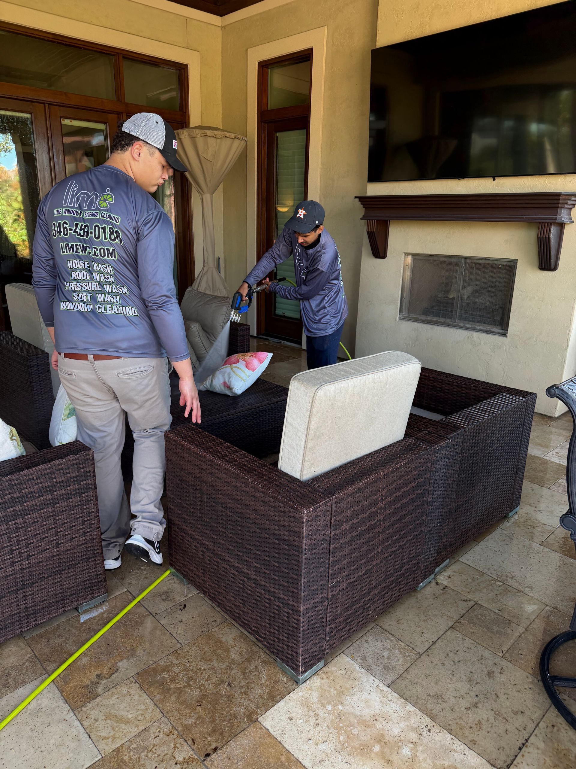 Two individuals in outdoor apparel clean and arrange dark wicker patio furniture on a stone terrace.