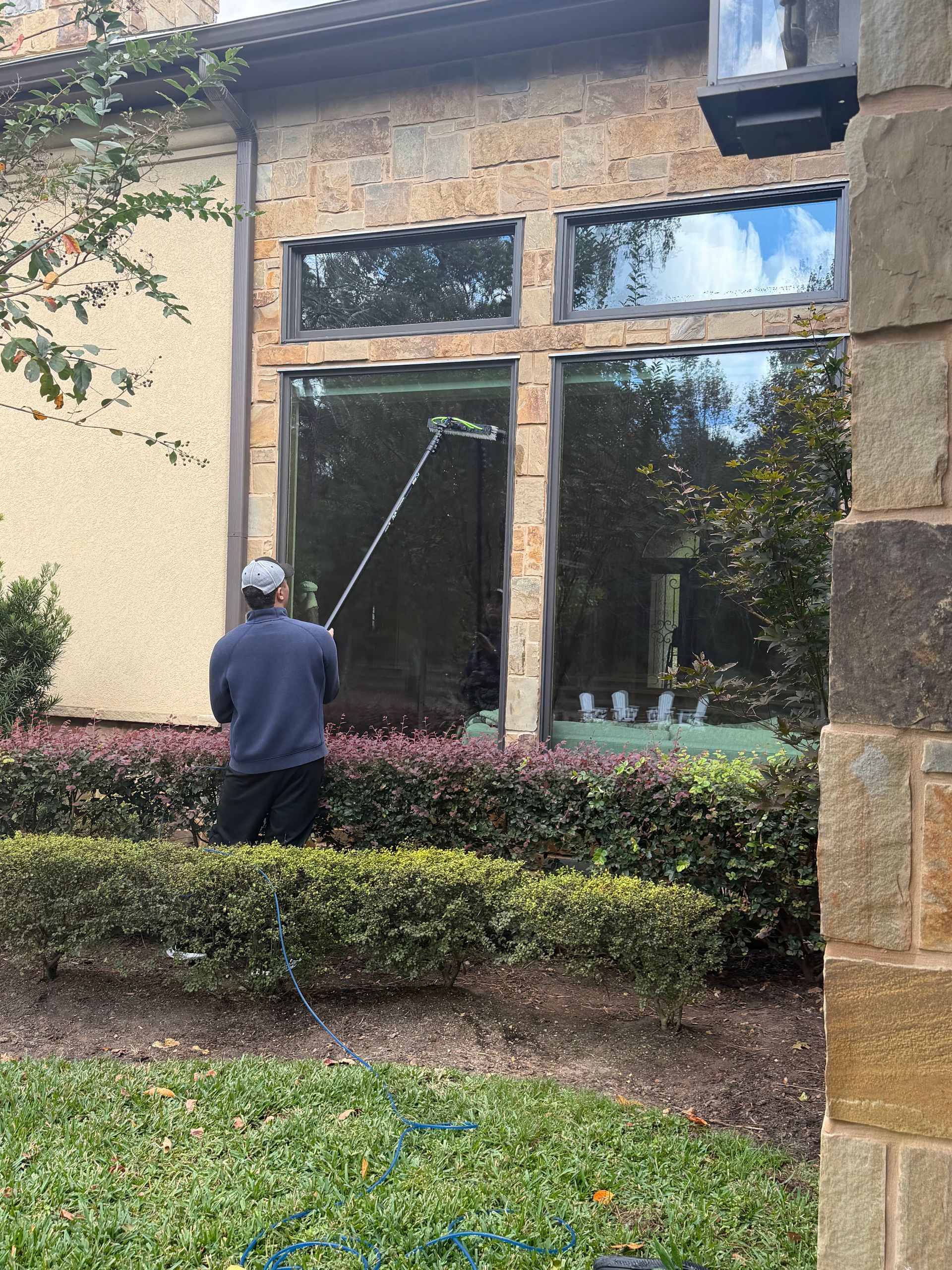 Person pressure-washes large windows on a stone house beside hedges and greenery.