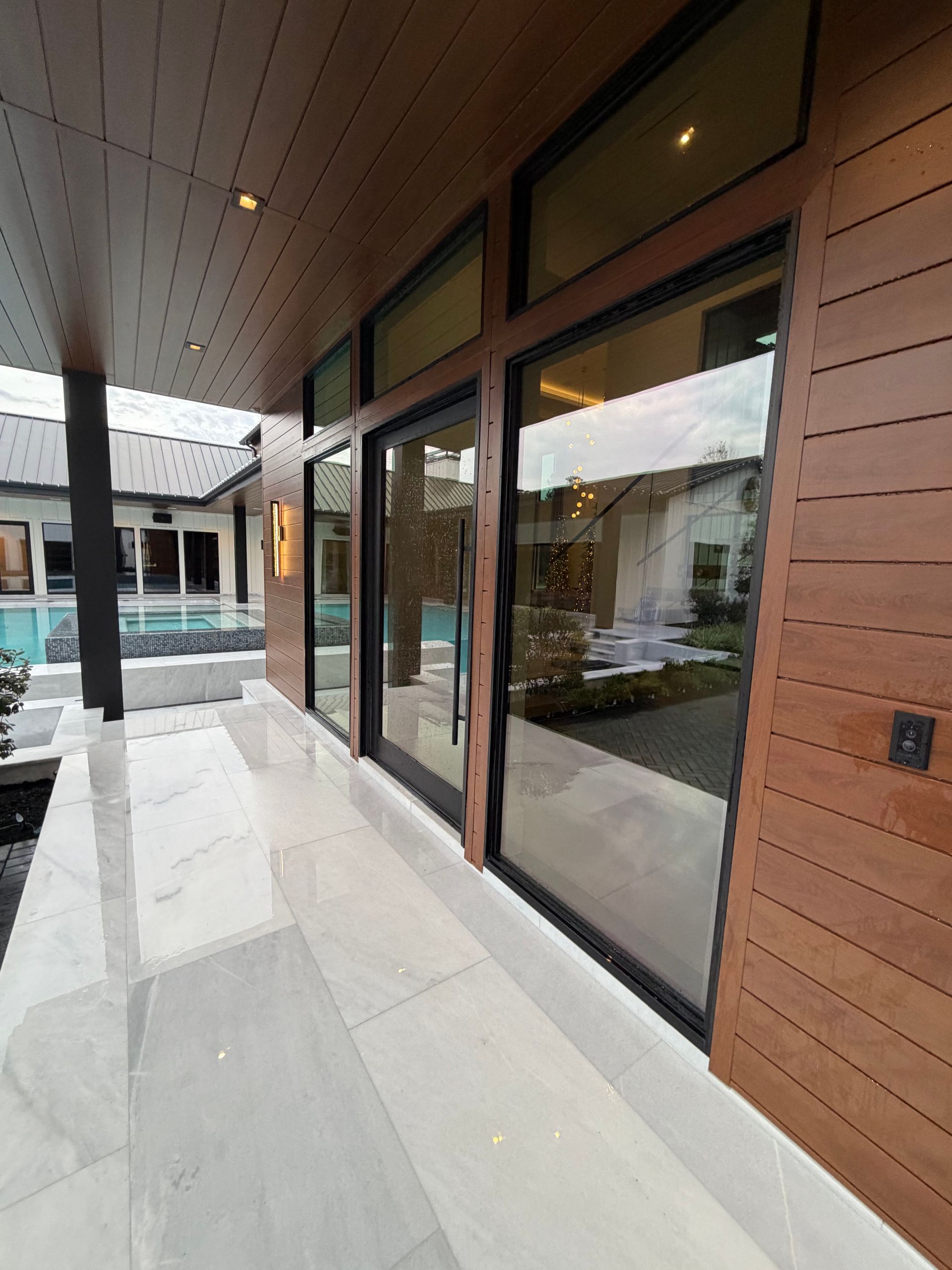 Covered modern patio with large glass doors and wood siding beside a pool