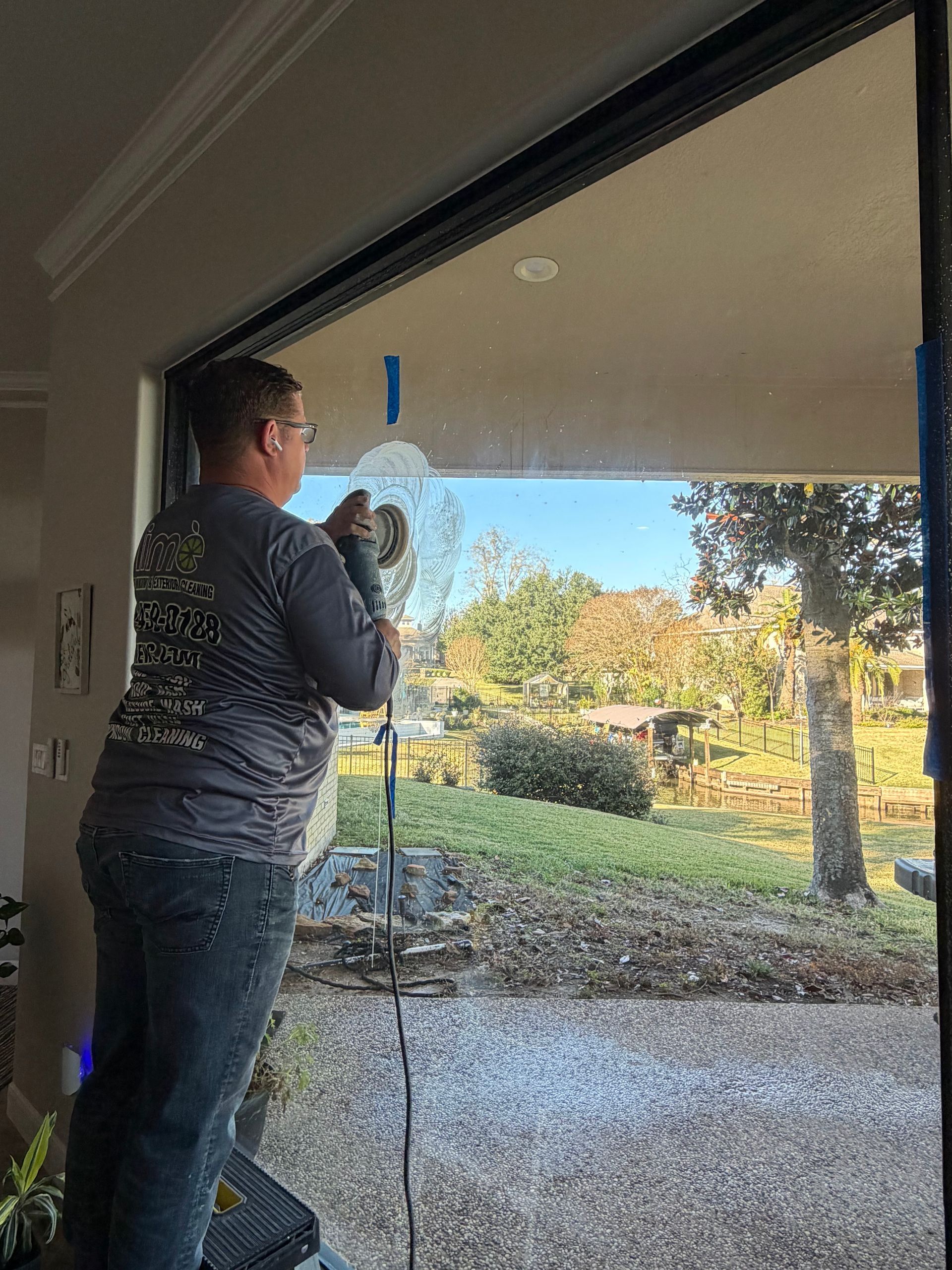Person cleaning a sliding glass door with a squeegee, standing inside a home facing a sunny yard.