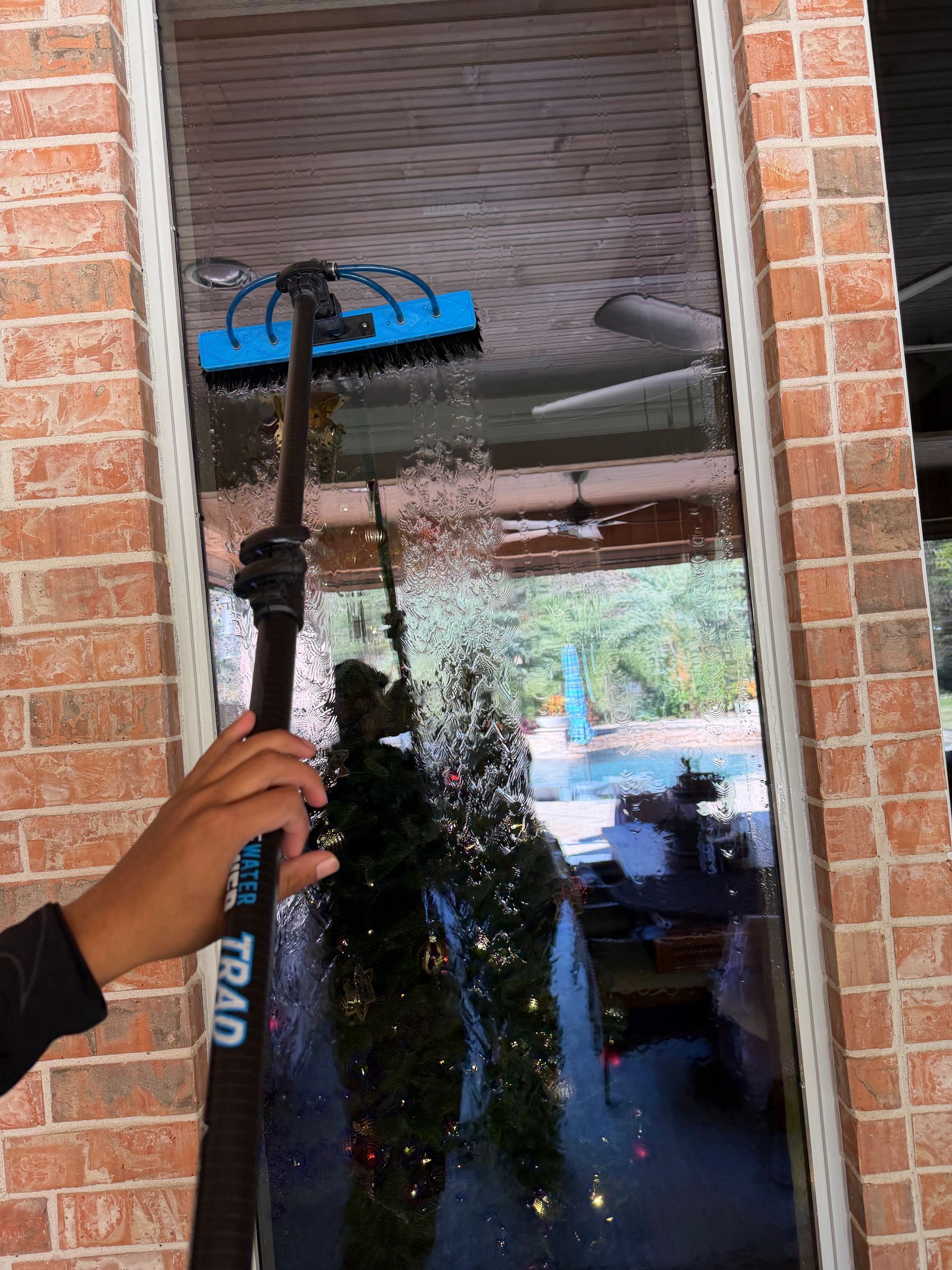 Hand holding a pressure washer spraying water on a glass window beside a brick wall