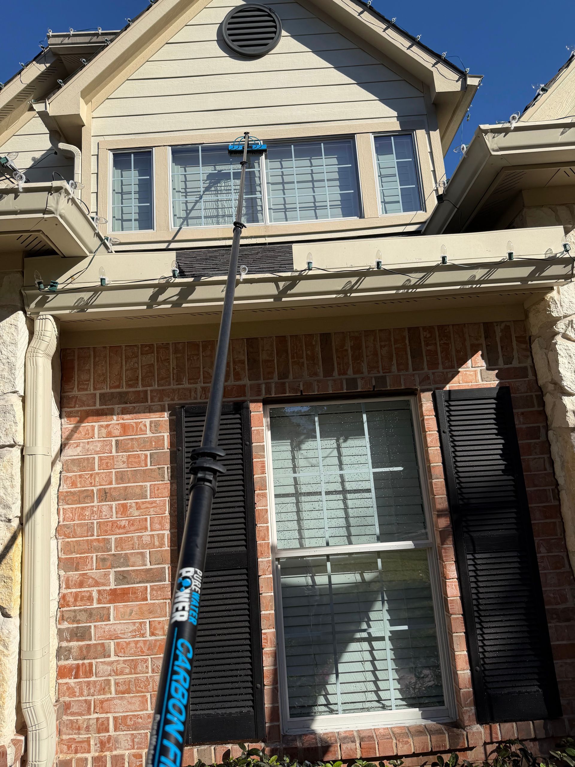 Person on ladder painting a two-story house with beige siding, brick facade, and black shutters