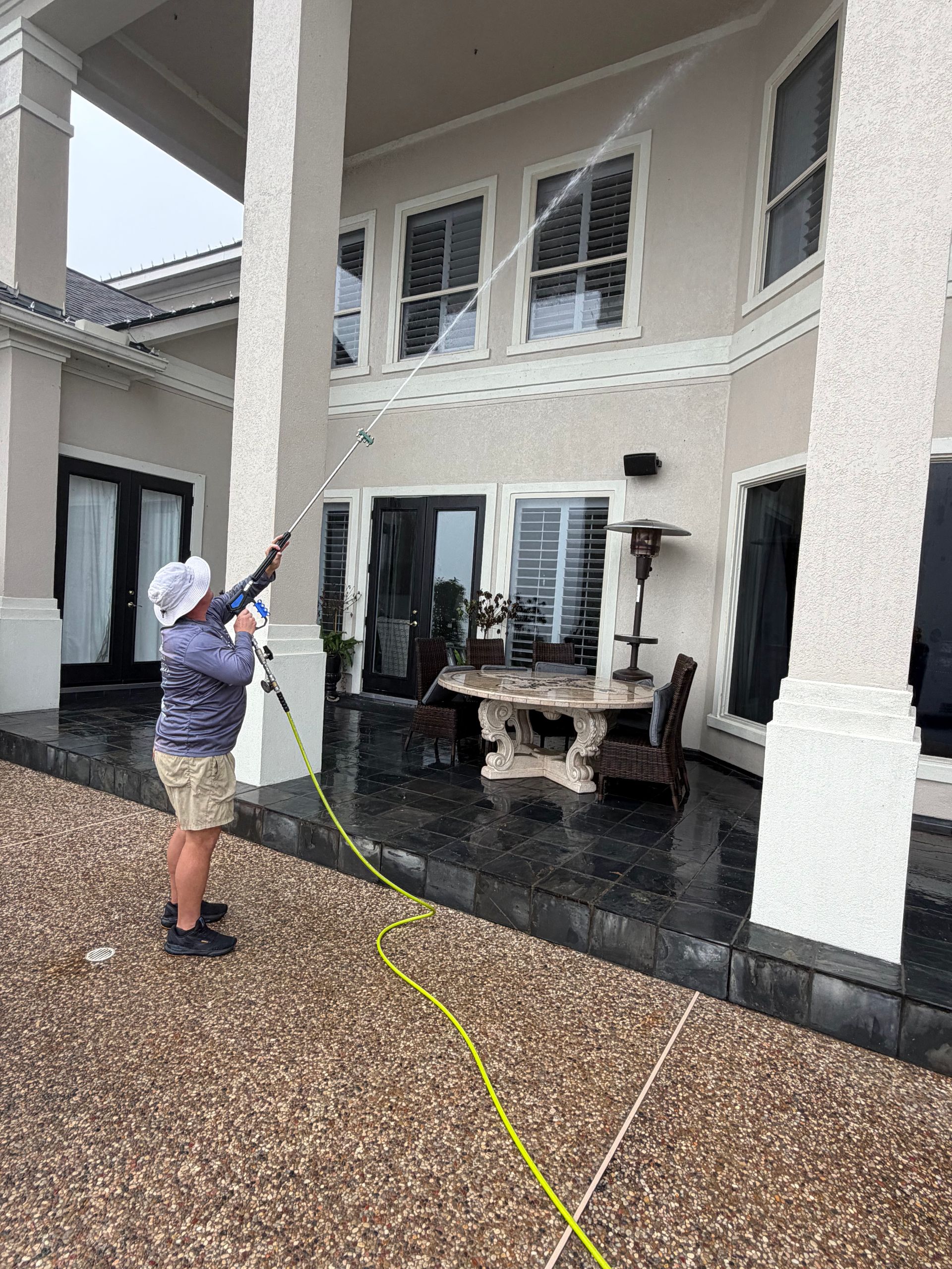 Person pressure-washing a covered patio outside a two-story house with white columns and black tile flooring