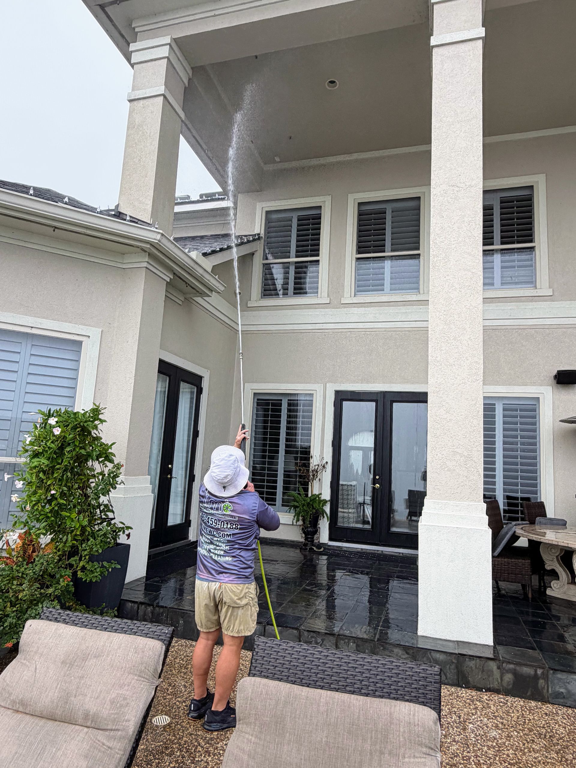 Person spraying a two-story house exterior, standing near a covered patio with white columns.