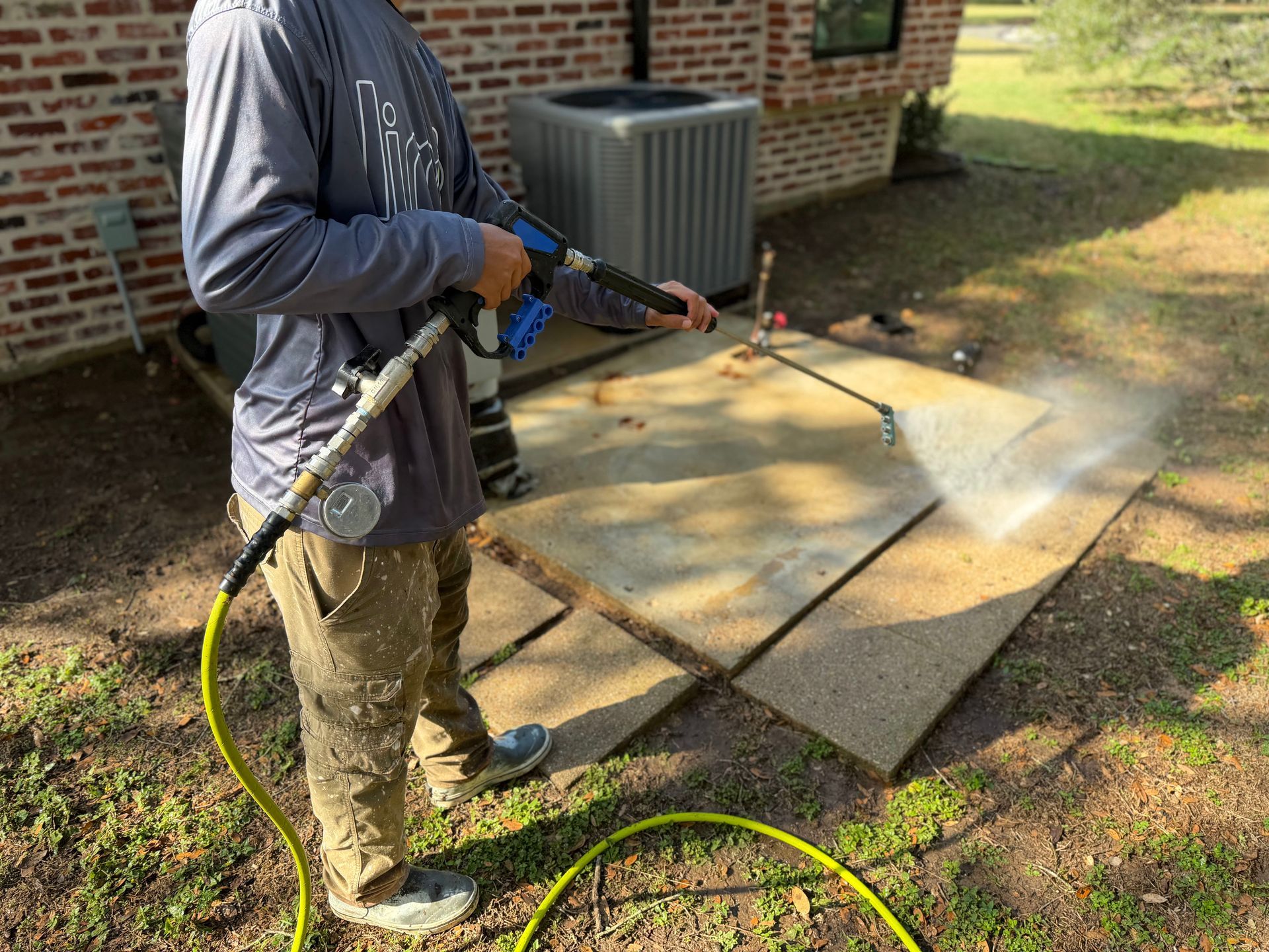 Person pressure washing patio slabs beside a brick house with a yellow hose.