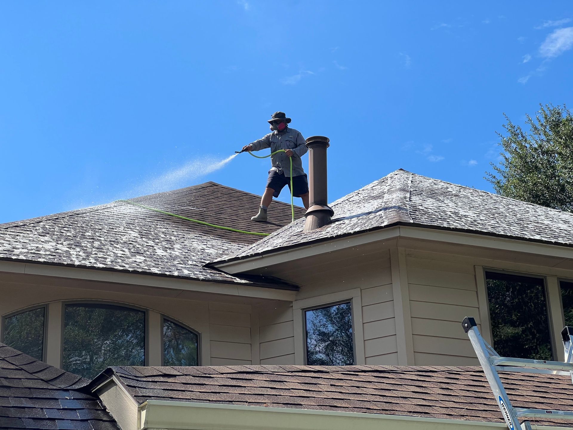 Worker repairing a shingled roof beside a chimney under a clear blue sky