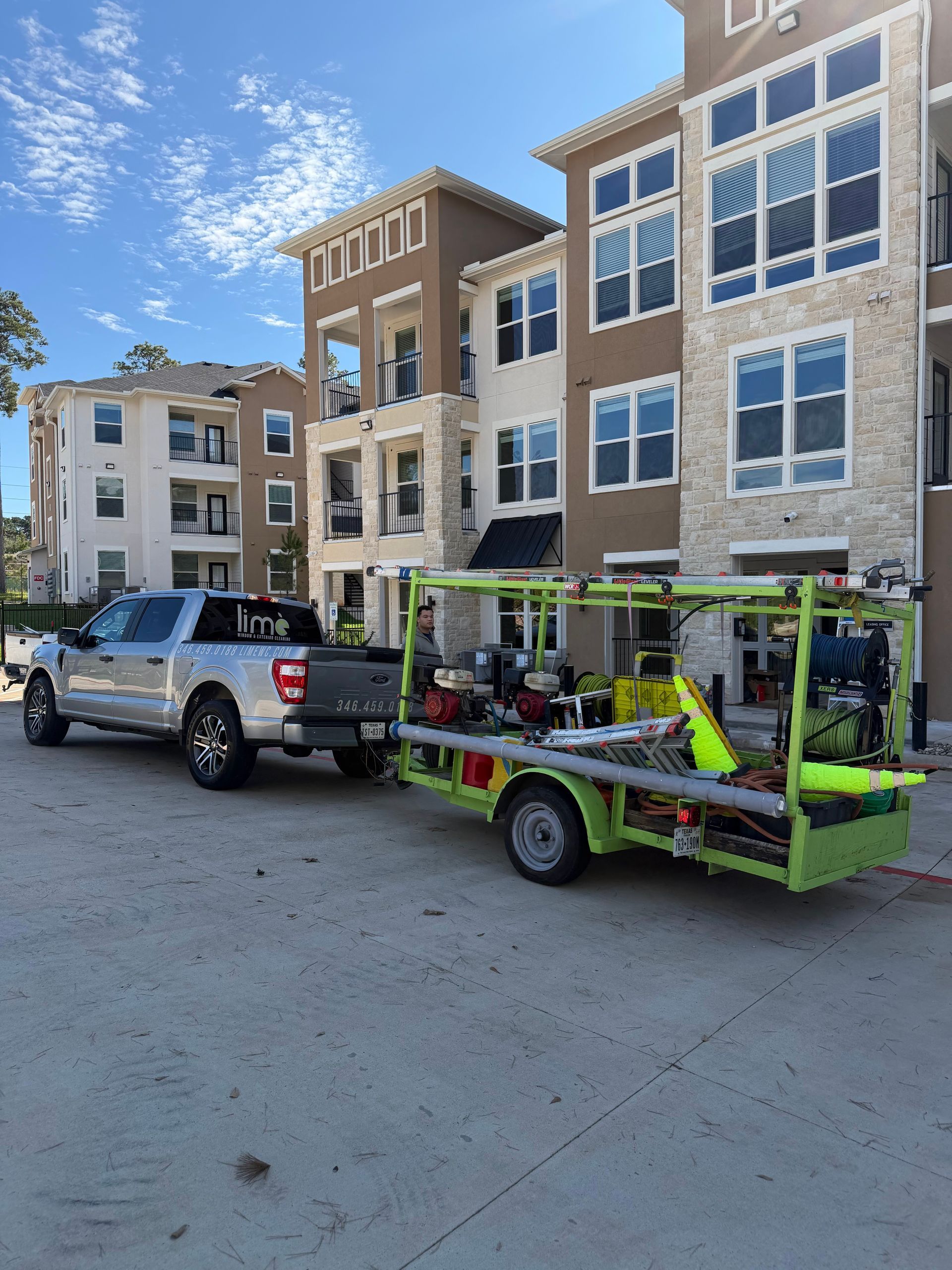 Pickup truck towing green utility trailer in front of apartment buildings on a sunny street.