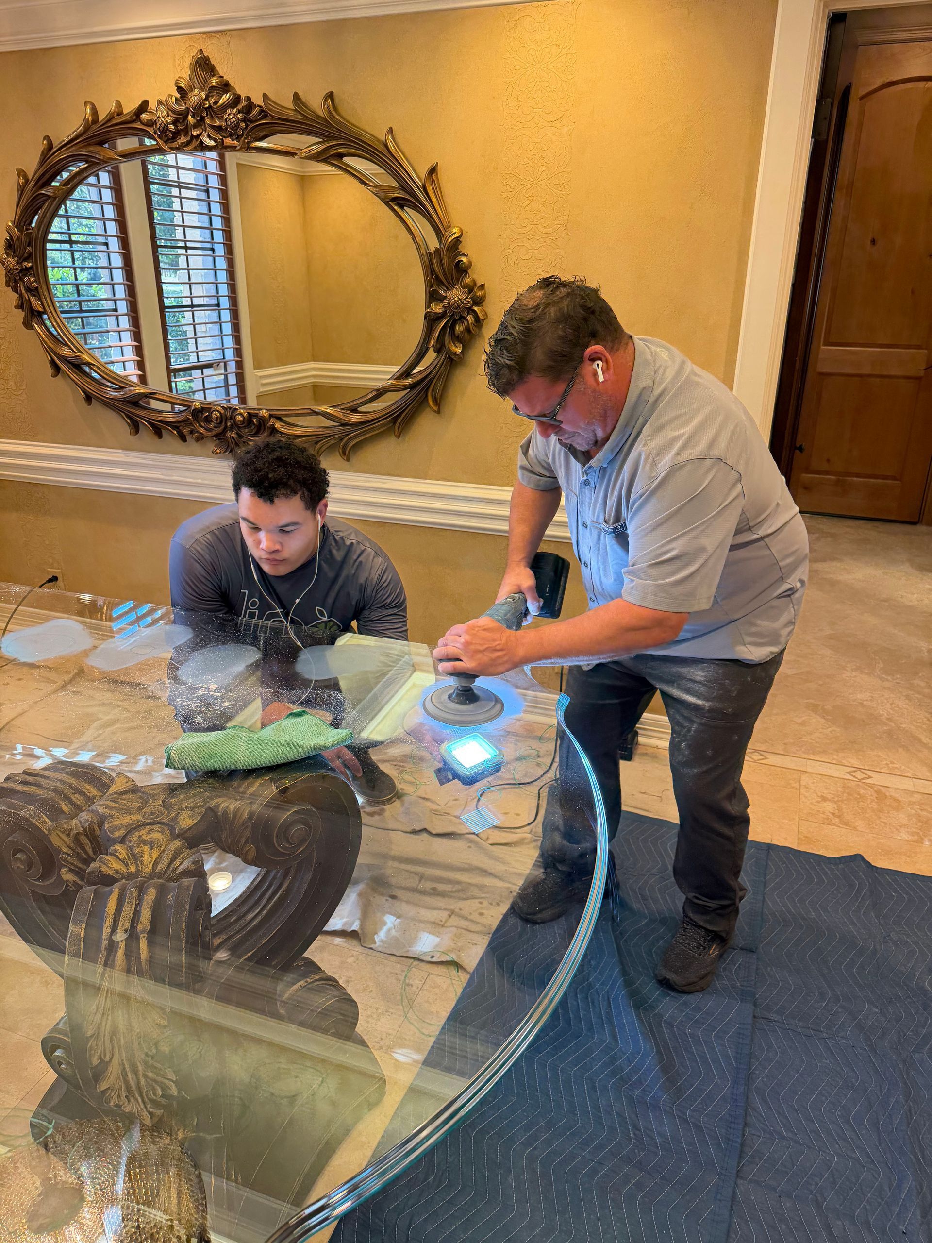 Two men examining a large glass tabletop in a living room beside a mirror and wooden door