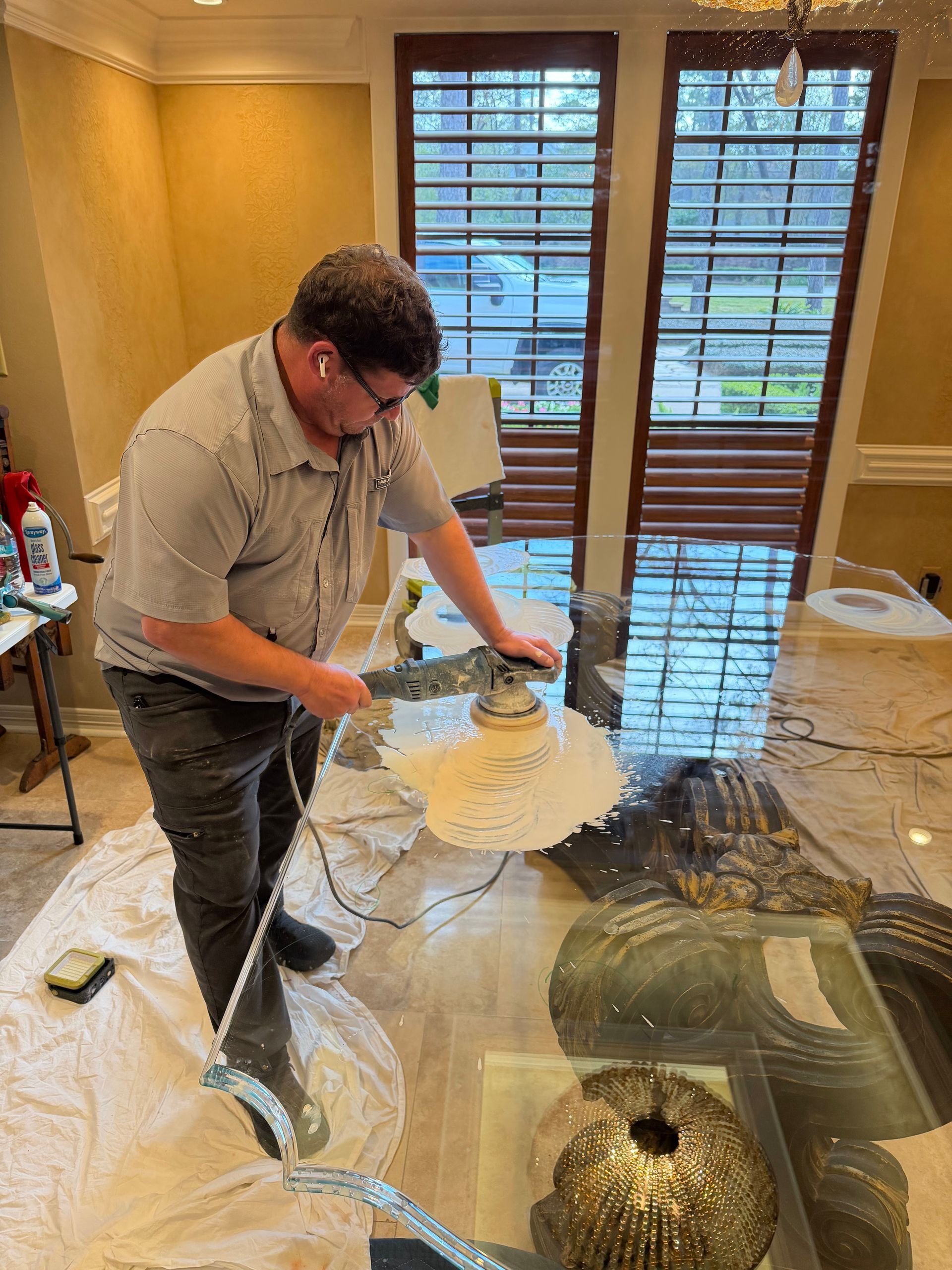 Man cutting protective film from a glass tabletop in a dining room with wooden shutters and a chandelier.