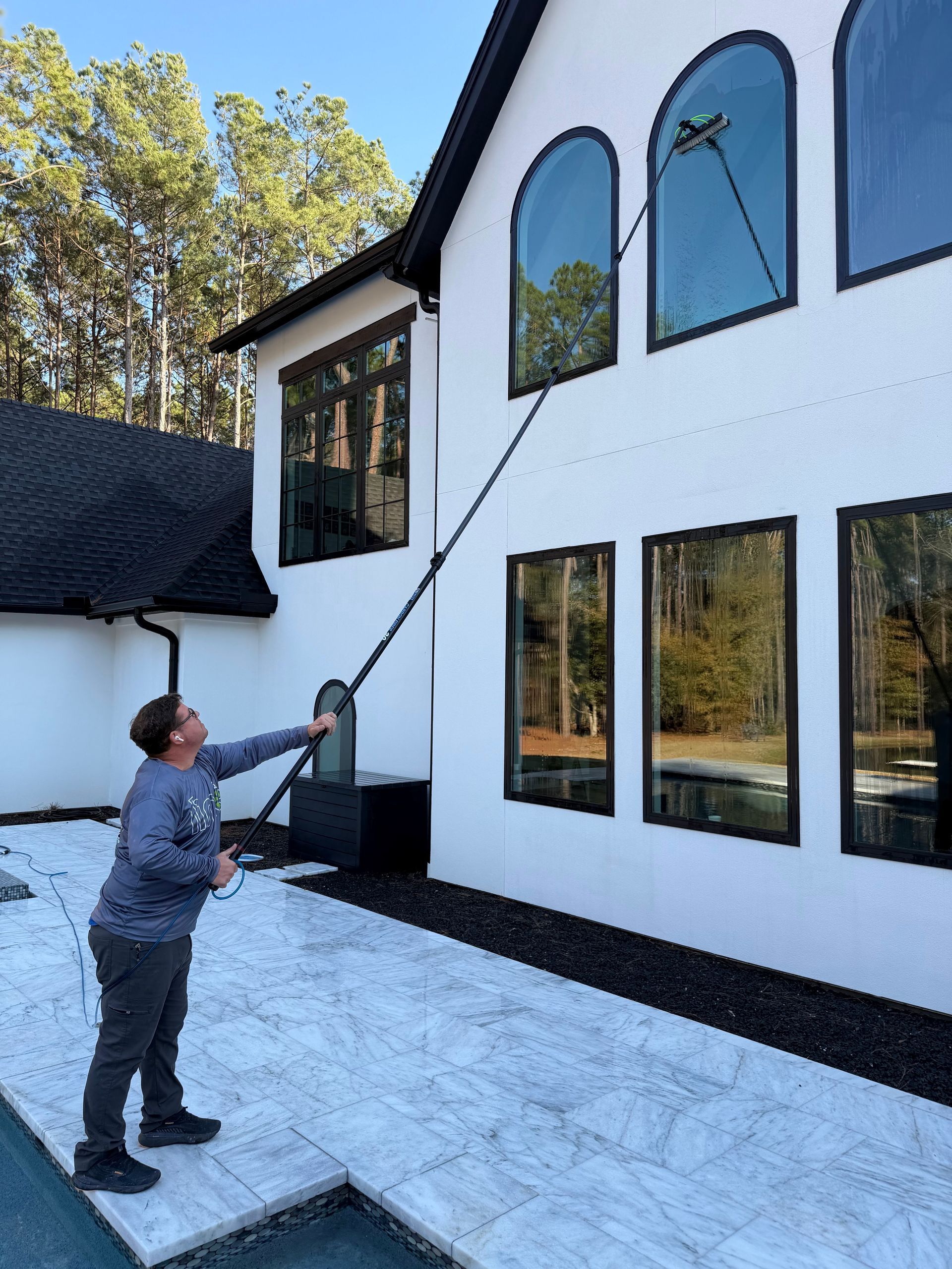 Person using a long pole to clean tall white house windows in snowy yard