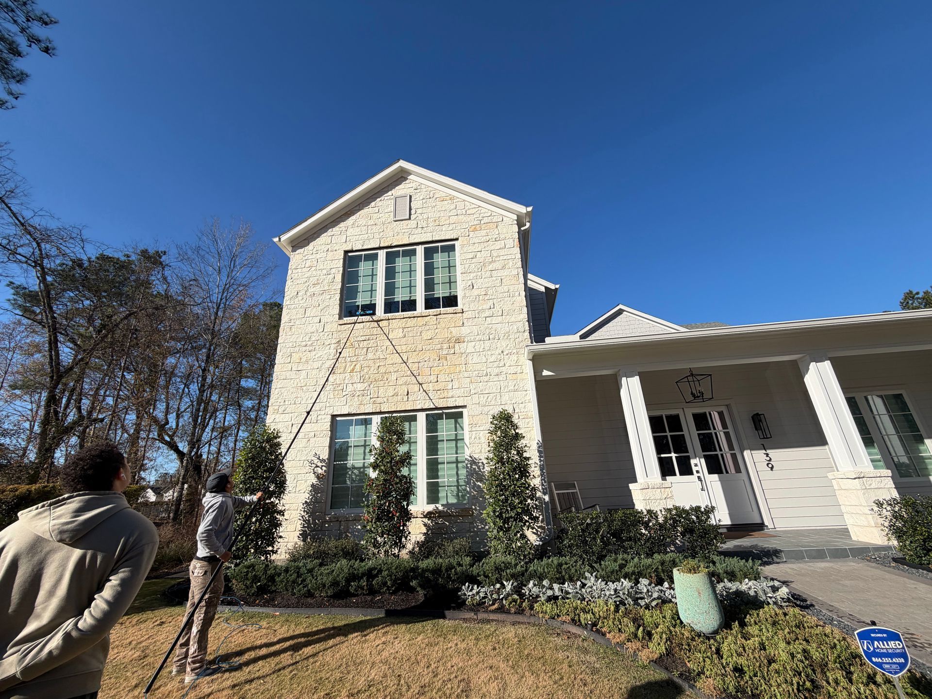 White stone house with green-trimmed windows and porch under a blue sky, with two people in the yard