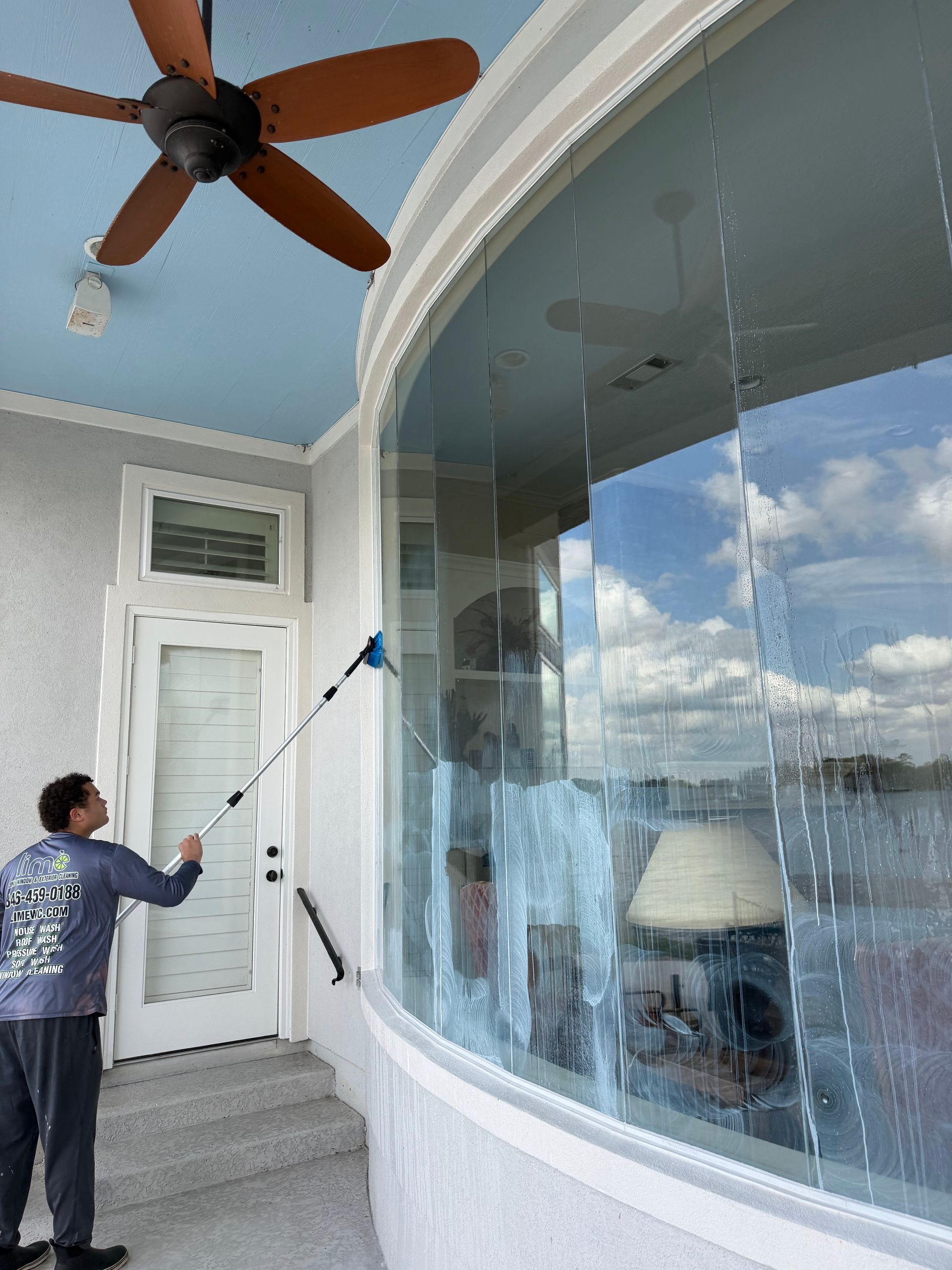 Person uses a long-handled brush to clean a large curved window on a porch.